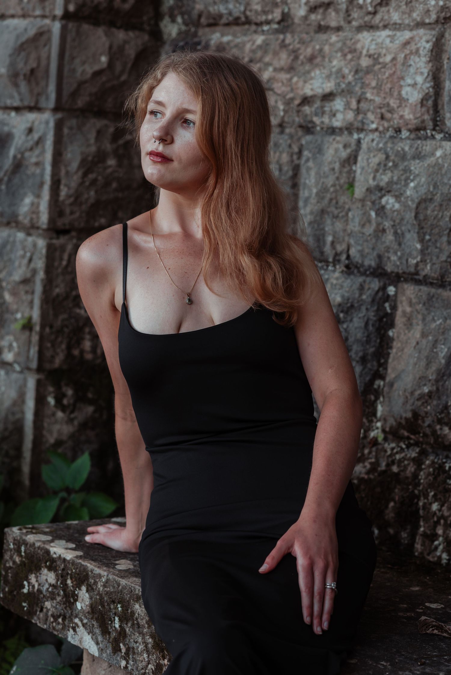 Young woman in black camisole leaning against an aged stone wall, looking thoughtfully to the side.
