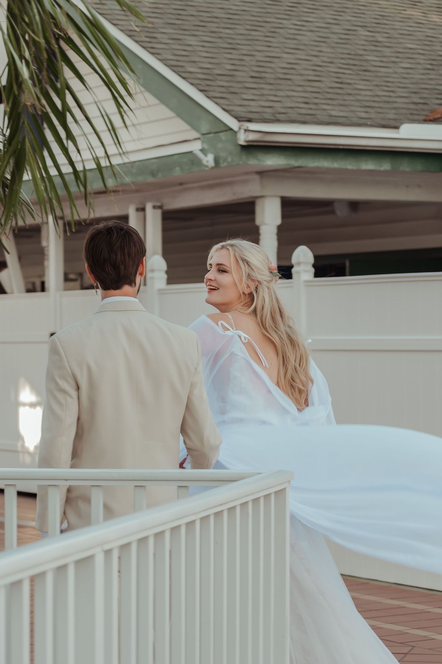 Smiling blonde bride in flowing white gown glancing back at groom on white railed walkway.