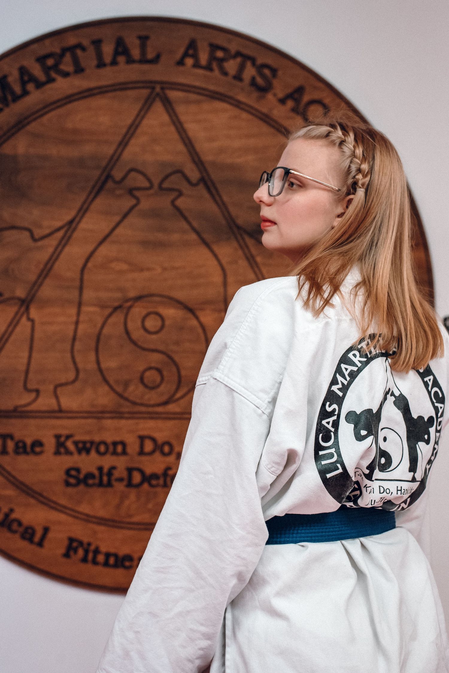 Female martial artist in white gi with blue belt posing before Lucas Martial Arts Academy sign.