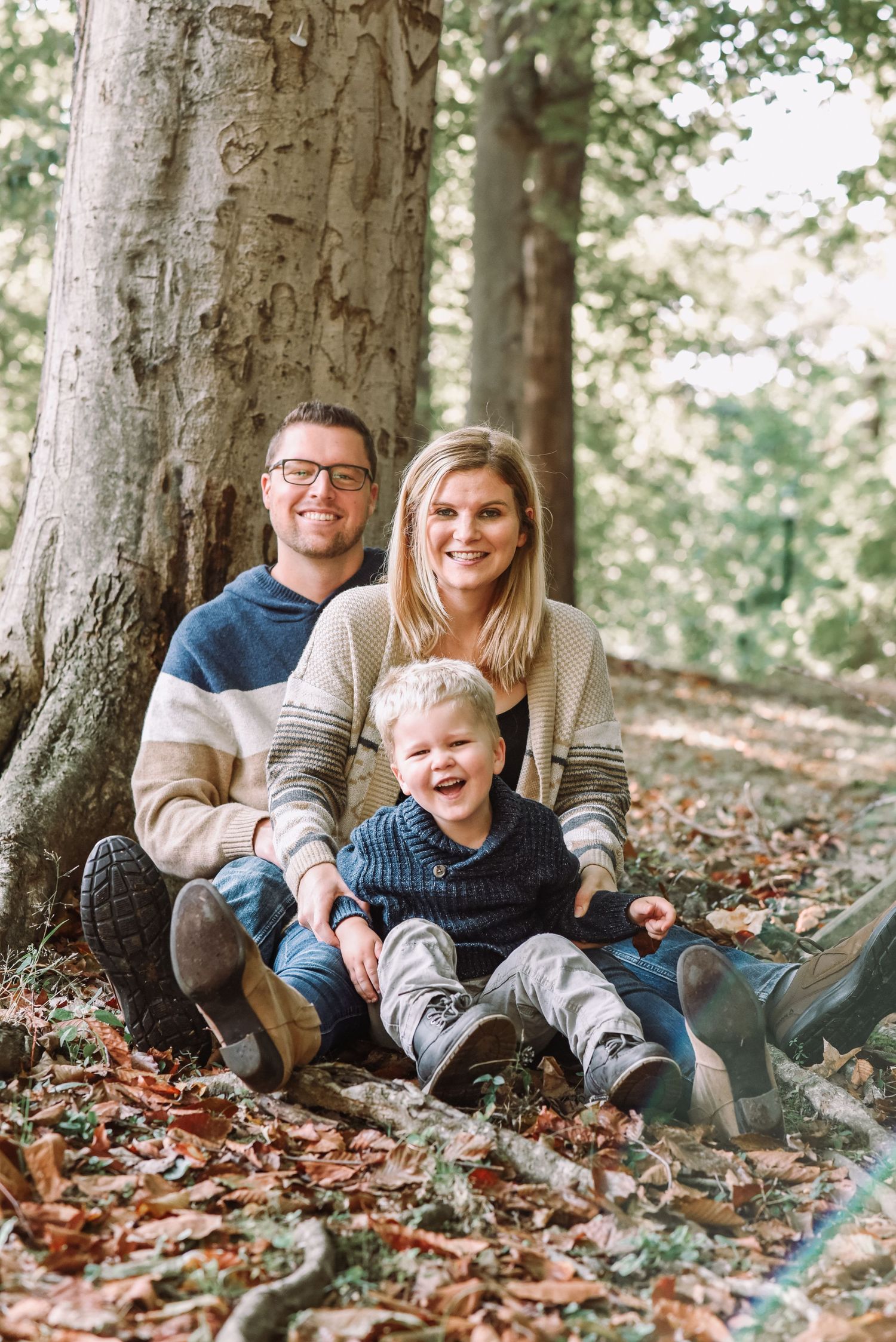 Smiling family of three sits together against a large tree trunk surrounded by autumn leaves outdoors.