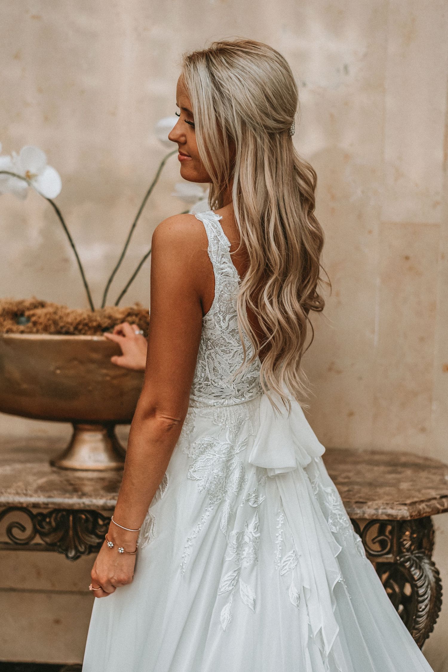 Blonde bride in sleeveless lace wedding gown with ruffle skirt poses beside ornate bronze planter.