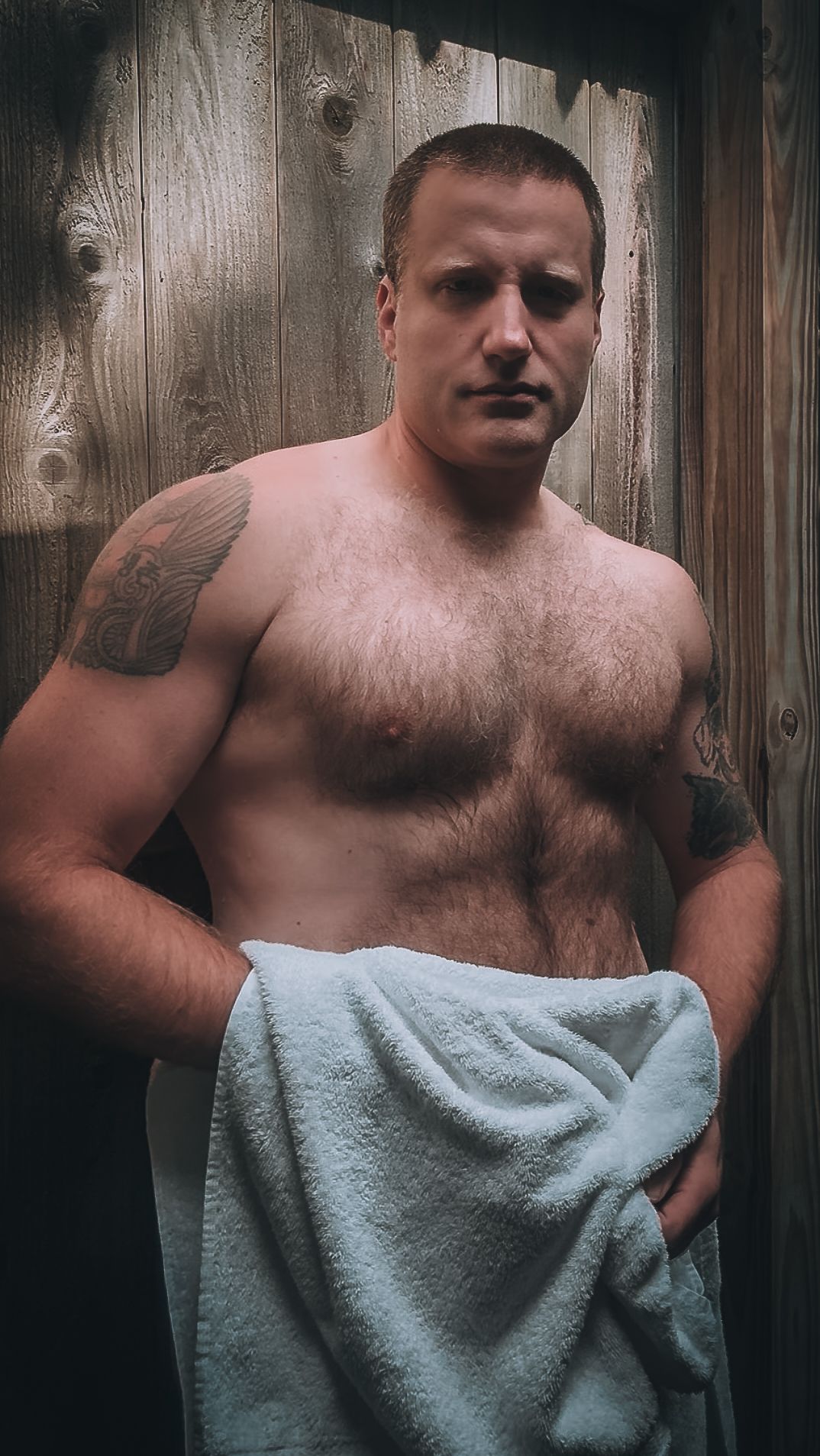 Shirtless muscular man with tattoo standing against wooden fence background.