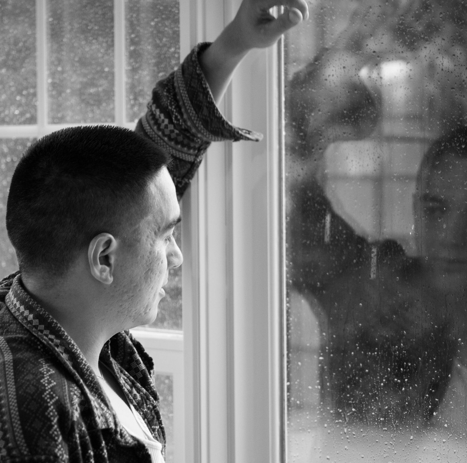 Black and white photo of a person looking through a rain-covered window.