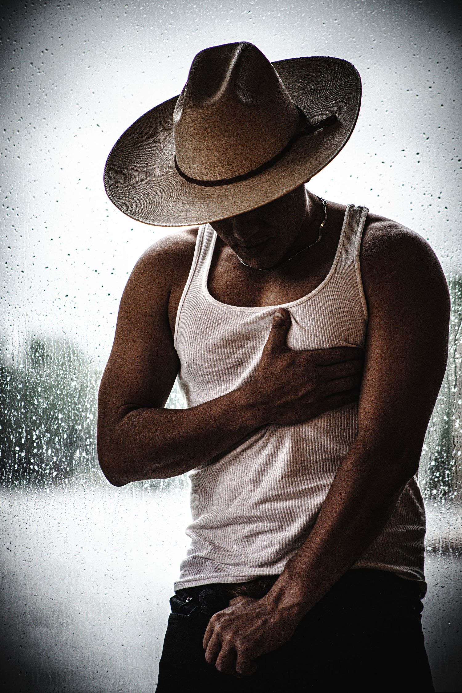 Moody portrait of a figure in a cowboy hat and tank top against a rainy window background.