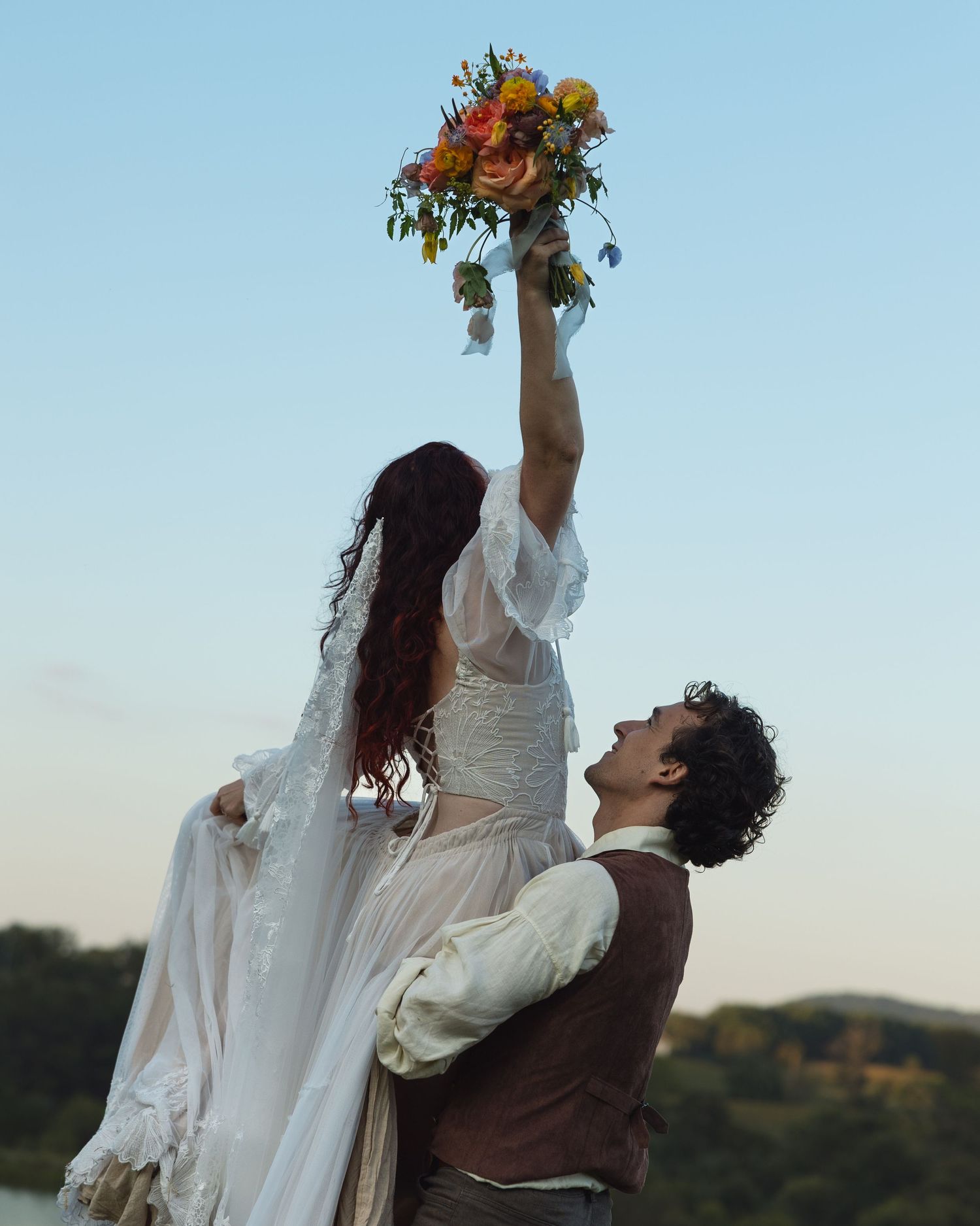 A romantic wedding couple poses outdoors at sunset, with the bride holding up a vibrant autumn bouquet.