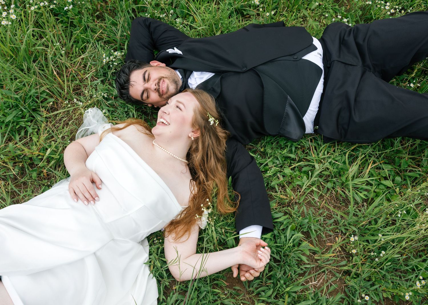 Bride and groom lying on green grass, laughing and holding hands in wedding attire.
