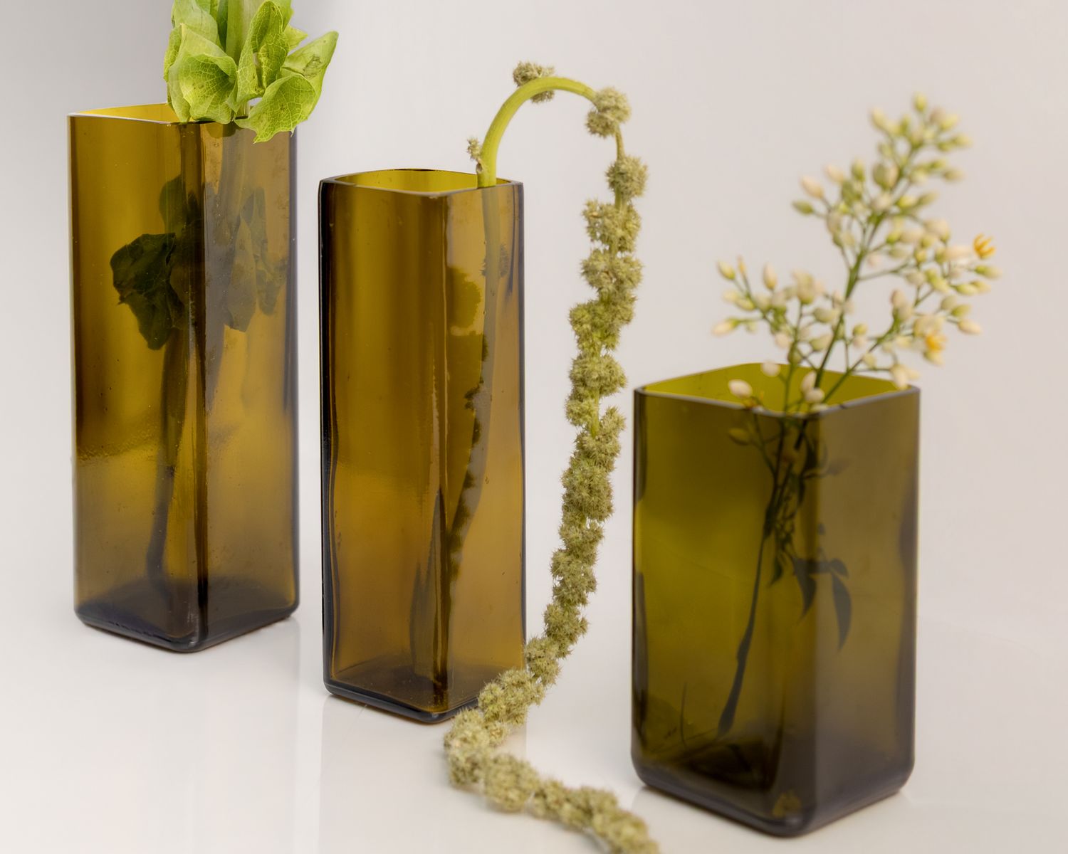 Three amber glass rectangular vases with dried and fresh botanical stems arranged on a white surface.