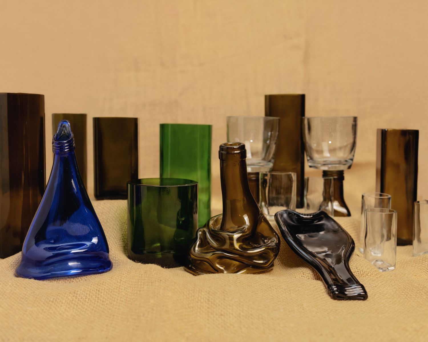 Collection of colorful glass vases, bottles, and drinkware in blue, green, and brown tones arranged on a beige surface.