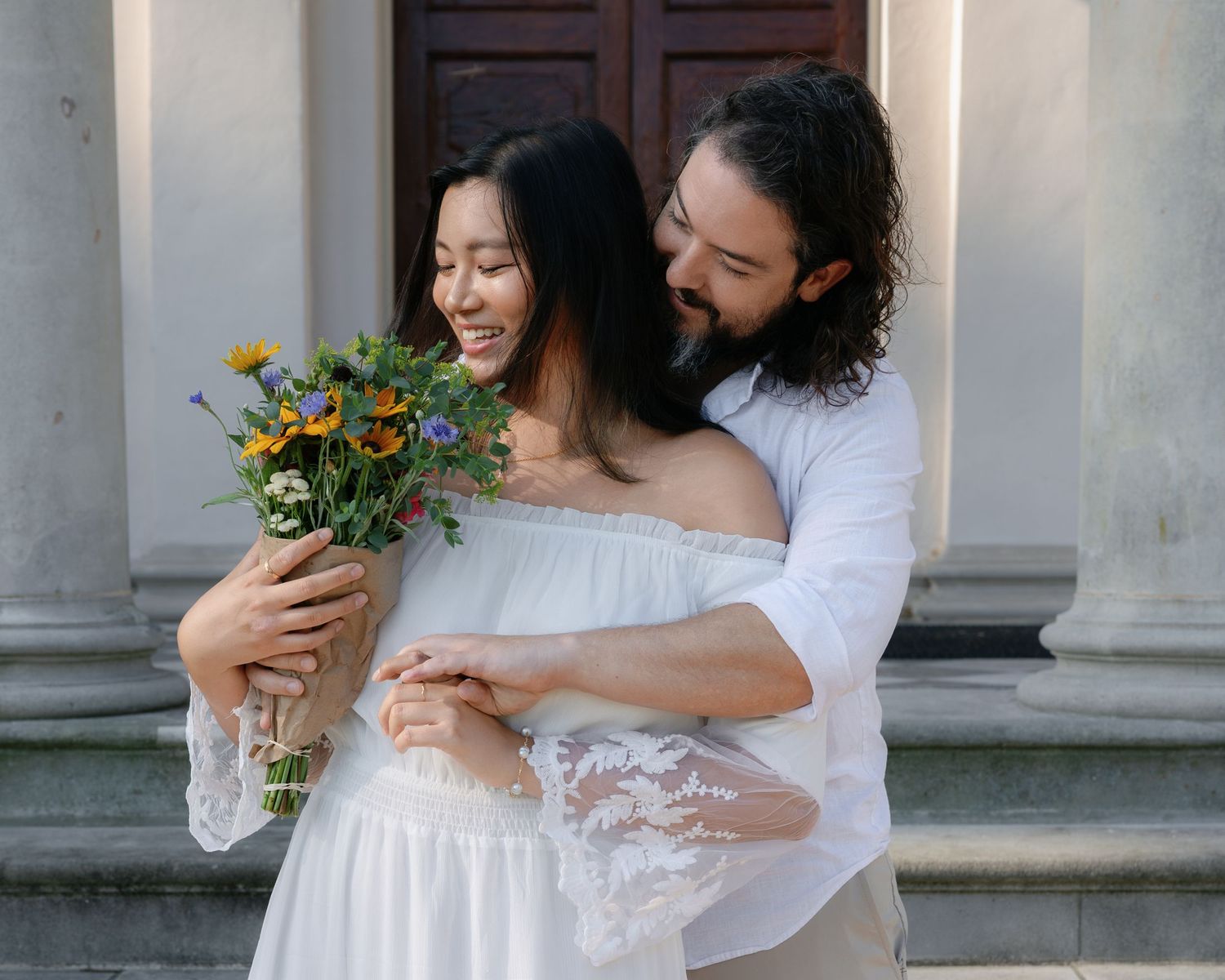 A romantic couple embraces outdoors while holding a colorful bouquet of wildflowers against a light stone backdrop.