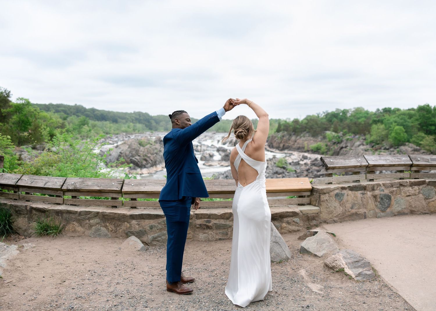 Groom in blue suit twirling bride in white backless gown at scenic overlook with waterfall and rocky landscape.