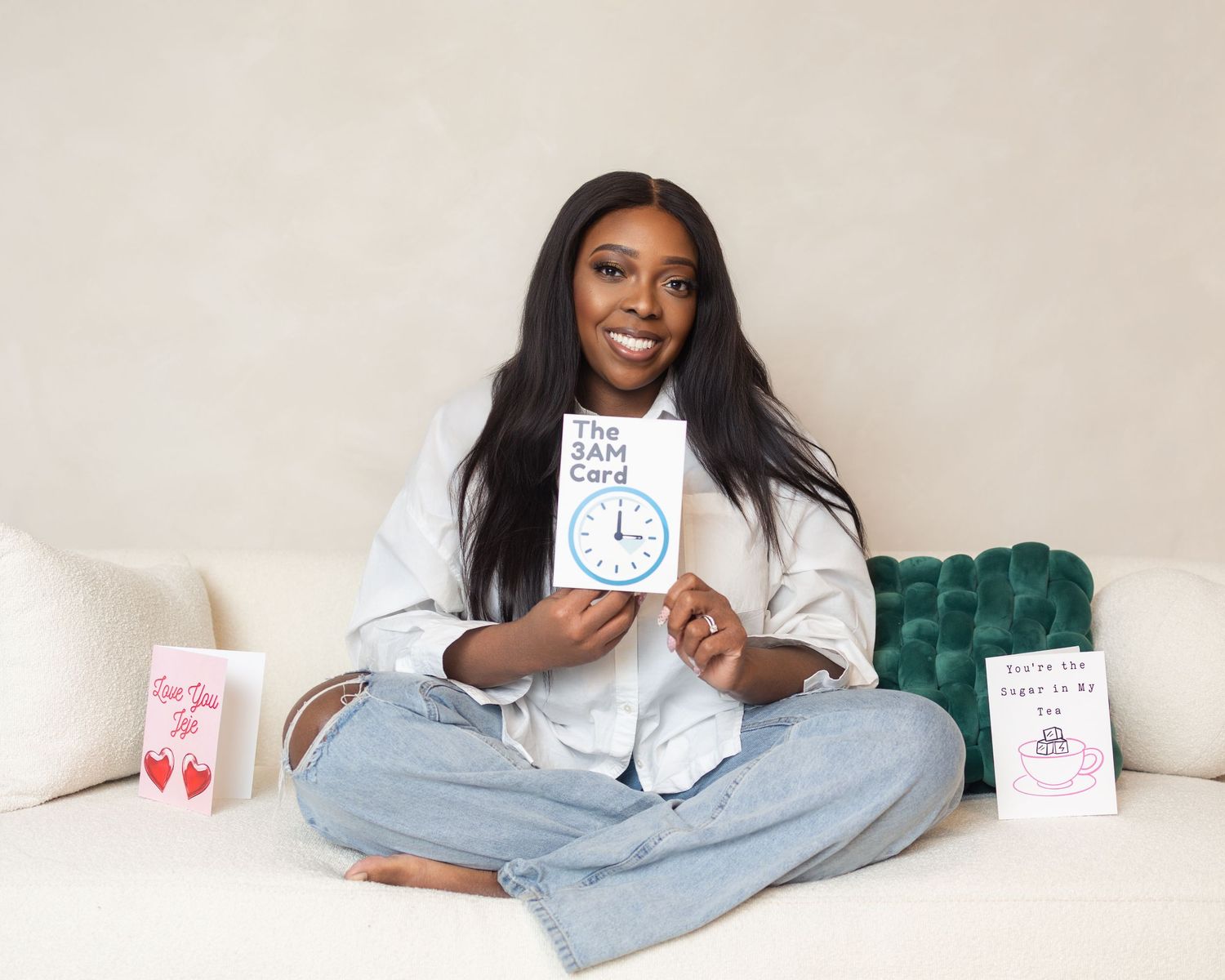 Woman sitting cross-legged on white sofa holding The 3AM Card, surrounded by greeting cards on display.