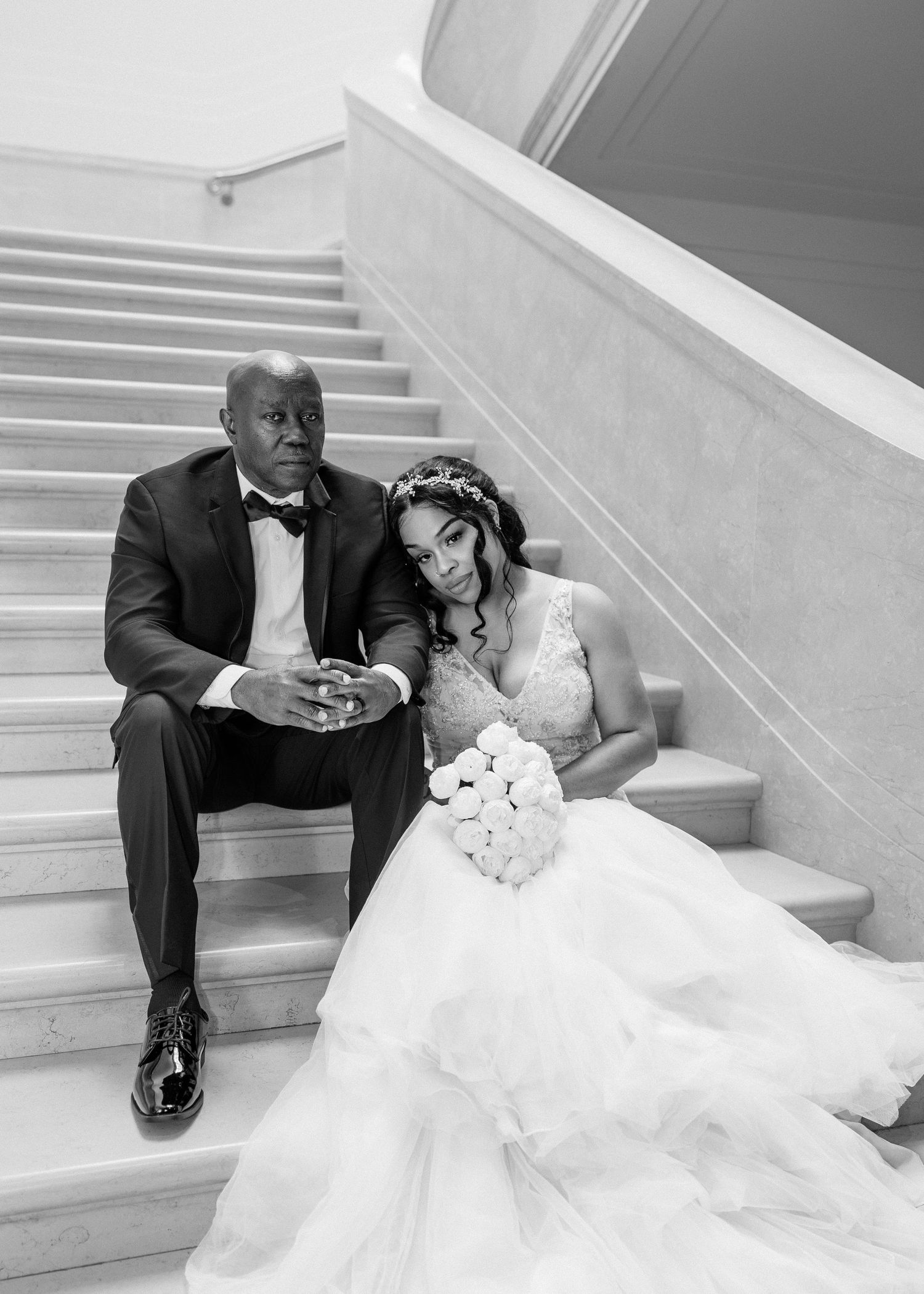 Elegant black and white wedding photo of bride and groom sitting on marble staircase, bride holding white rose bouquet.