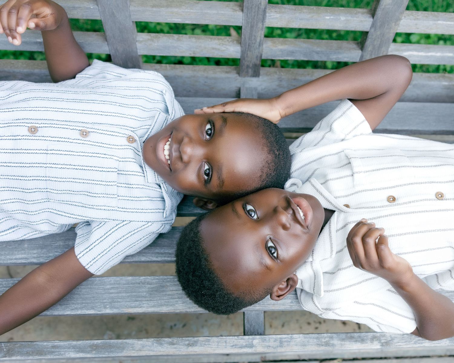 Two young boys in striped shirts lying on a wooden bench, smiling and looking up at the camera.