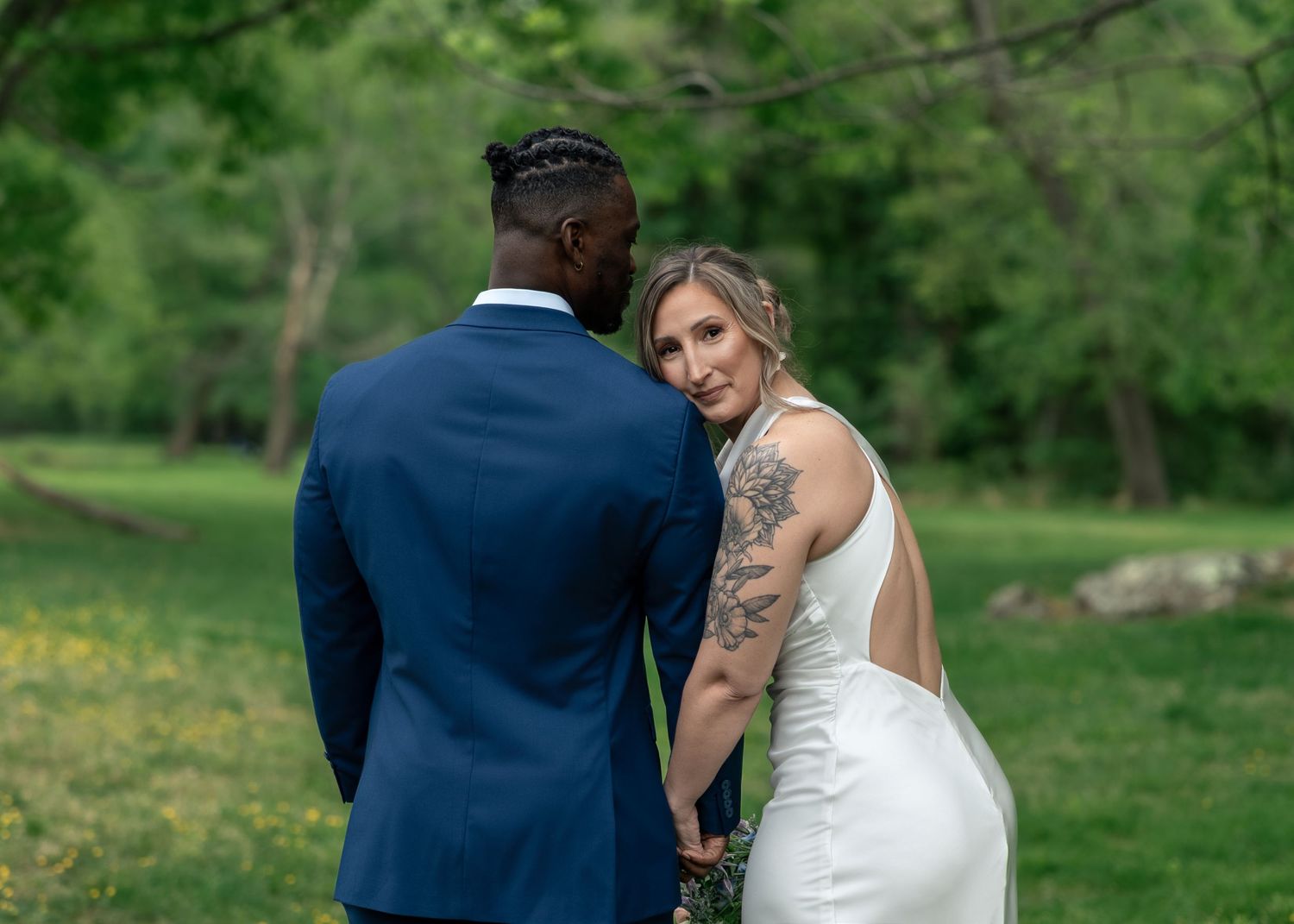 Bride with tattoo in backless white gown holds hands with groom in navy suit outdoors in lush green park setting.