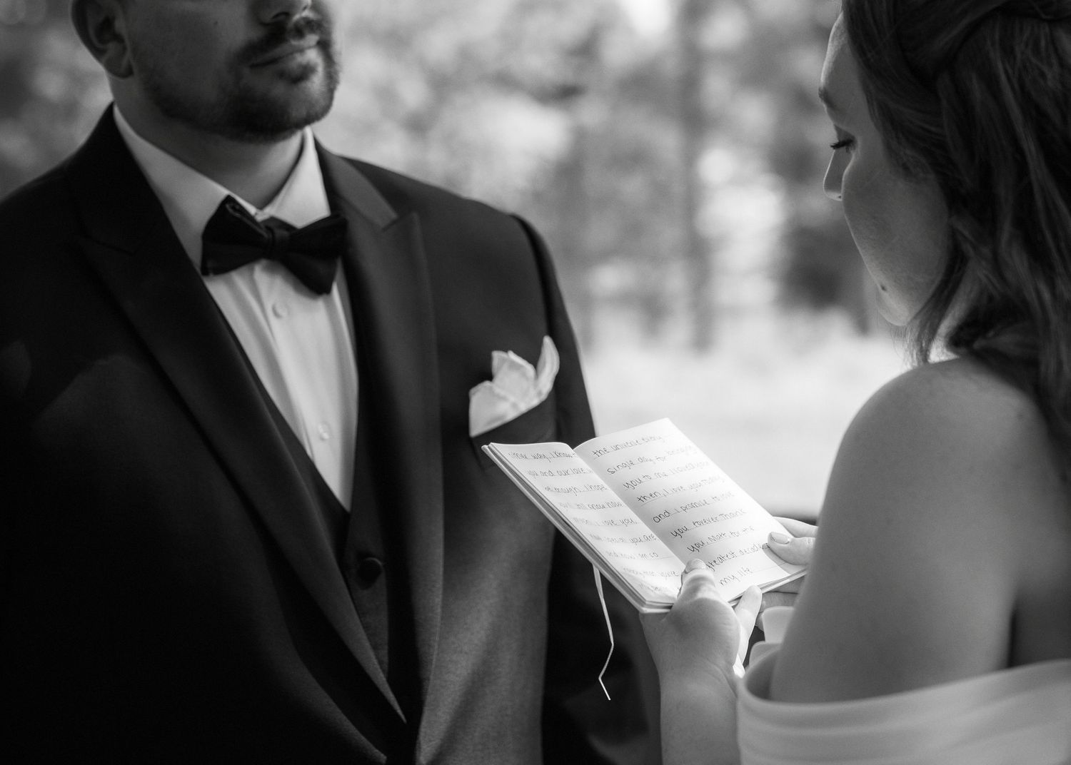 Bride reads wedding vows from paper to groom in tuxedo during intimate outdoor ceremony, black and white photo.