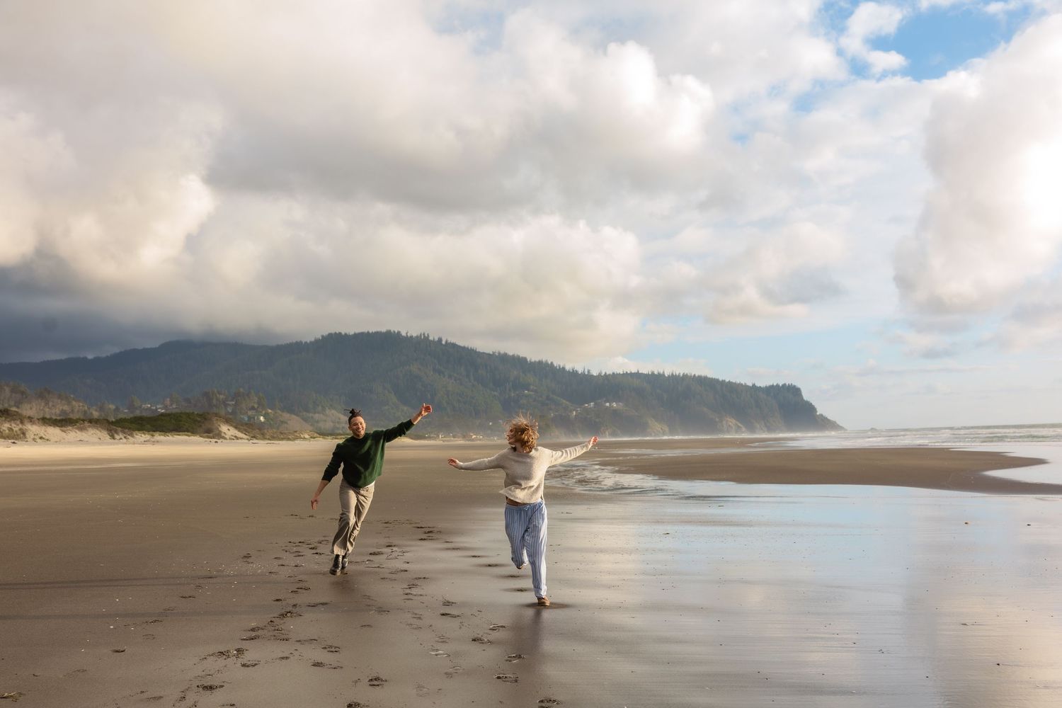 Couple joyfully running on a sandy beach with dramatic cloudy skies and green hills in the background.