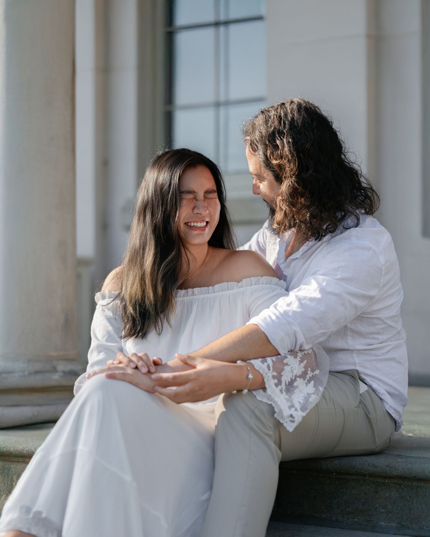 Two people in white clothing share intimate moments while sitting together in a bright interior space.