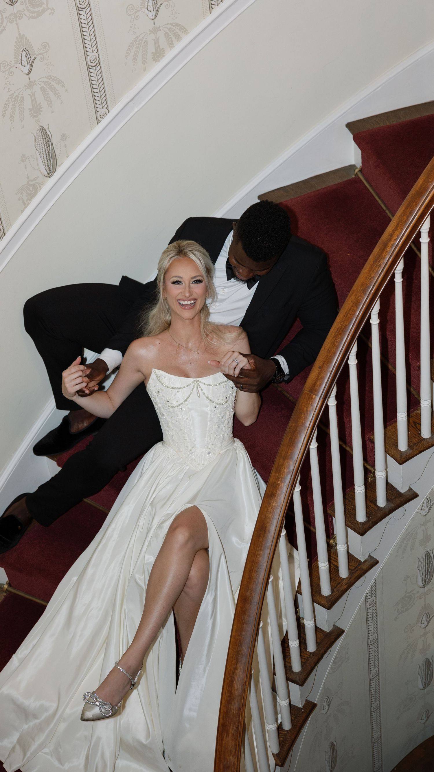 A couple poses on an elegant curved staircase with white walls and red carpeting.