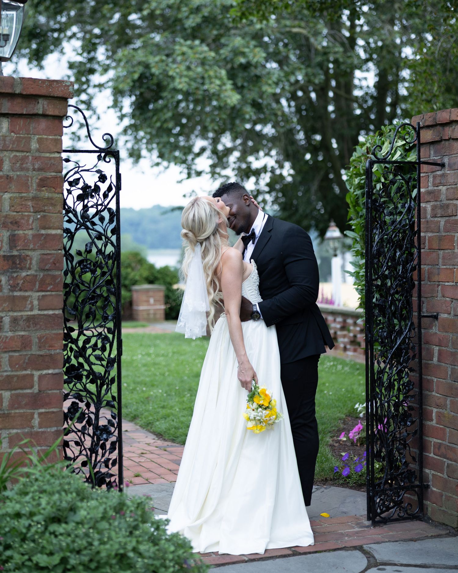 A couple shares a romantic kiss at an outdoor wedding venue framed by ornate wrought iron gates and brick pillars.