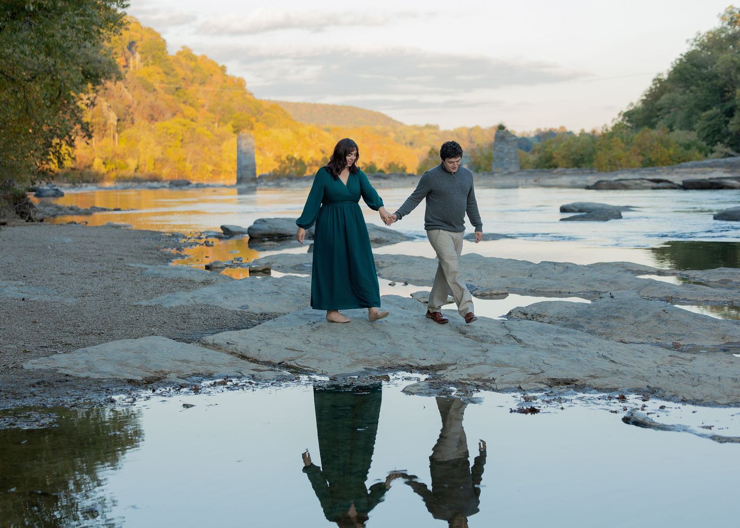 Two people walk together on rocky riverbank with autumn foliage and reflective puddles during cloudy day.