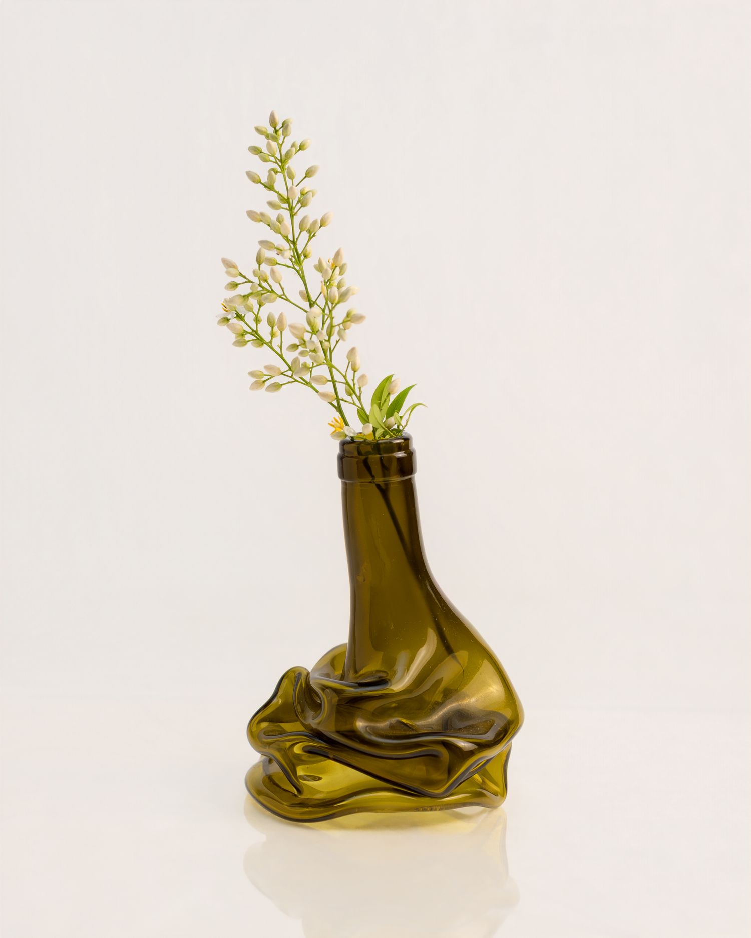 Olive green crumpled glass bottle vase holding a delicate white flowering branch on white background.