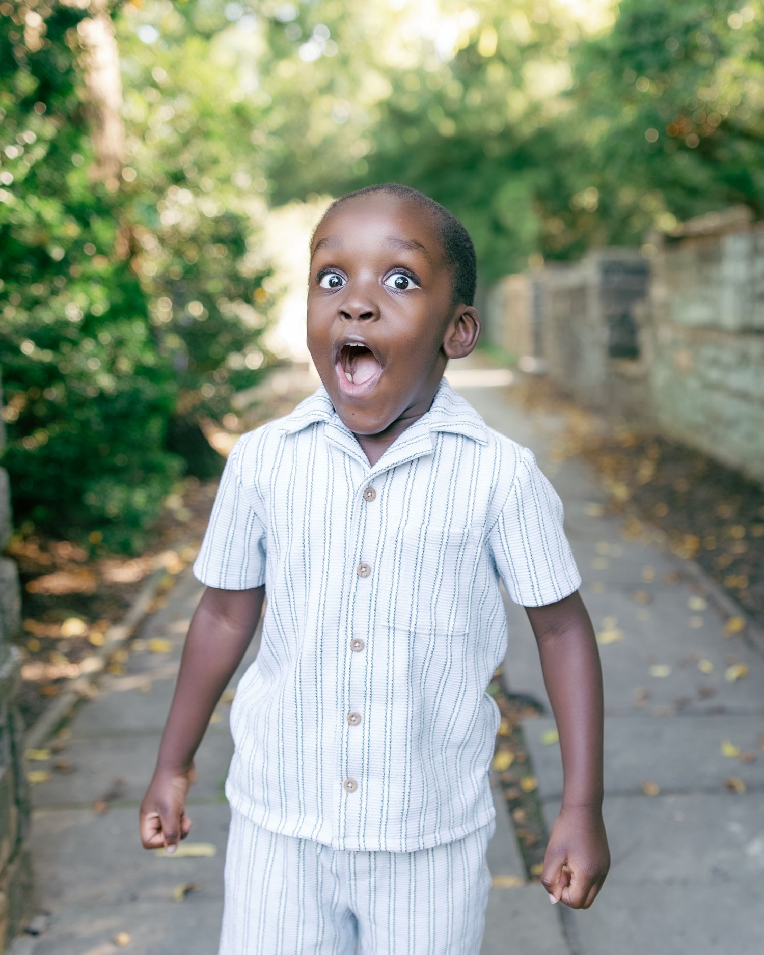 A young Black boy in a striped short-sleeve outfit stands on a path, mouth open wide in surprise, with green trees behind.