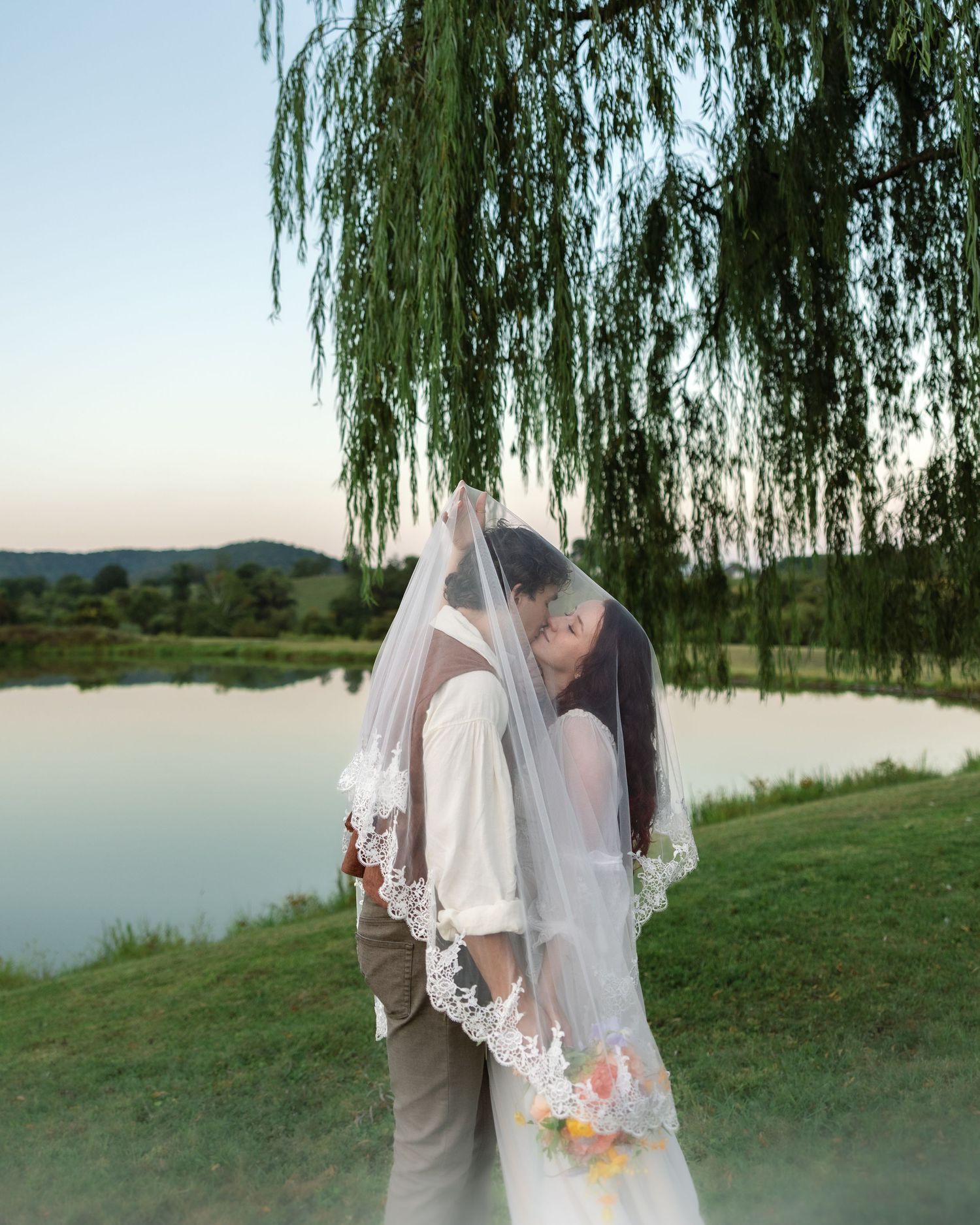 A romantic wedding couple embraces under a weeping willow tree beside a tranquil lake at sunset.