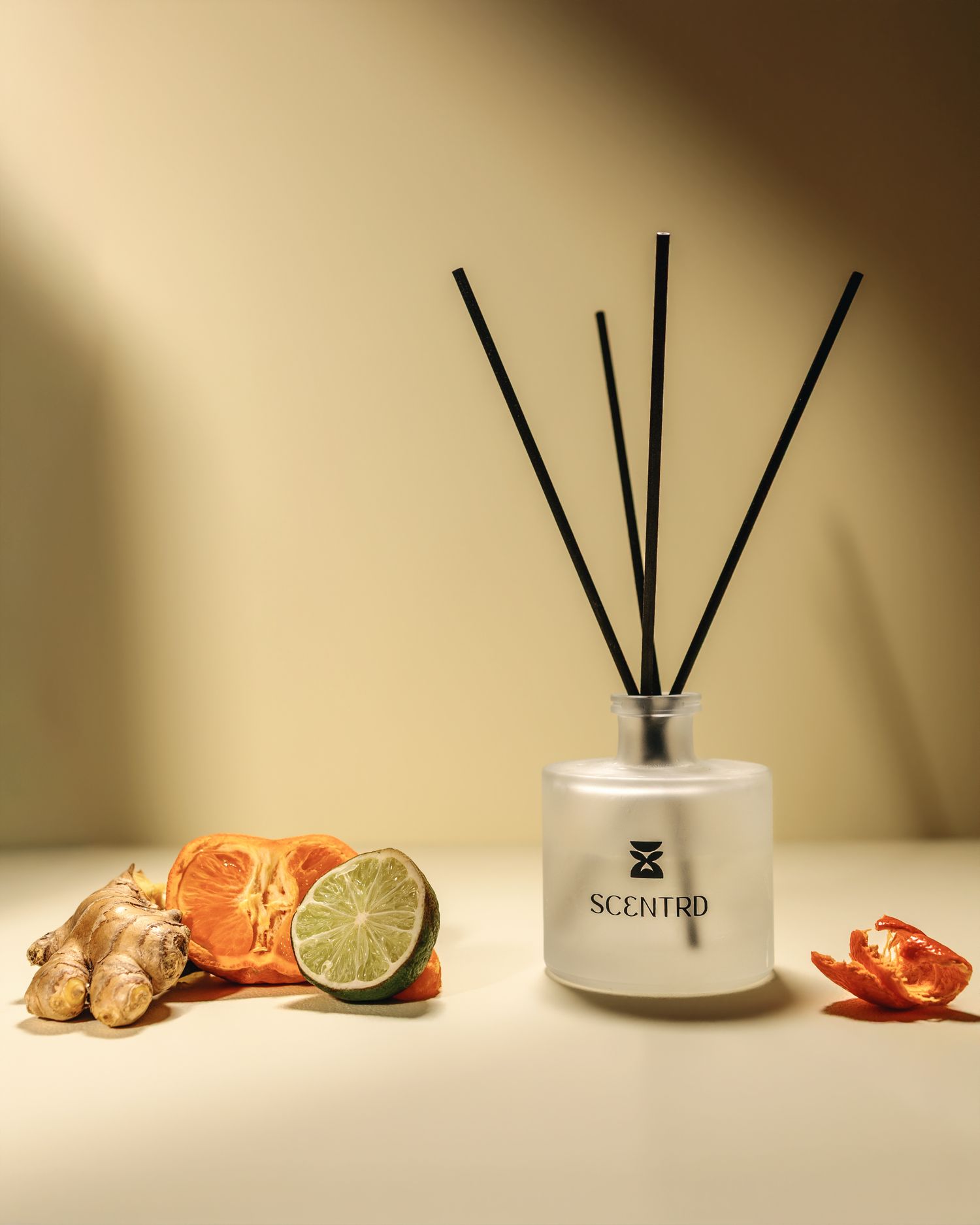 Scentrd reed diffuser in frosted glass with black reeds, surrounded by ginger, lime, and orange on a beige background.