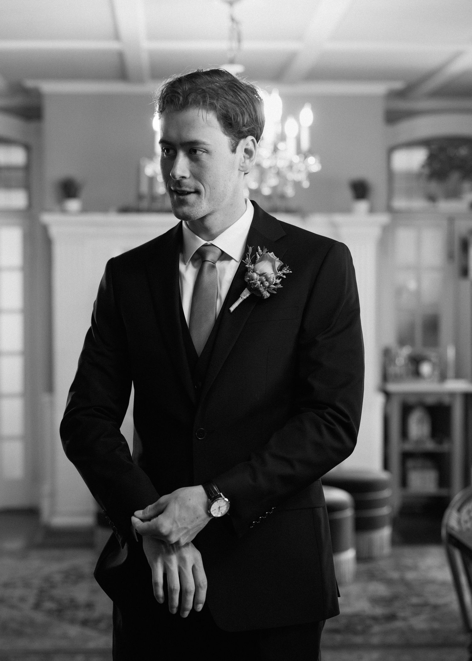Black and white portrait of a groom in a dark suit with boutonniere and tie, adjusting his cuffs in an elegant venue.
