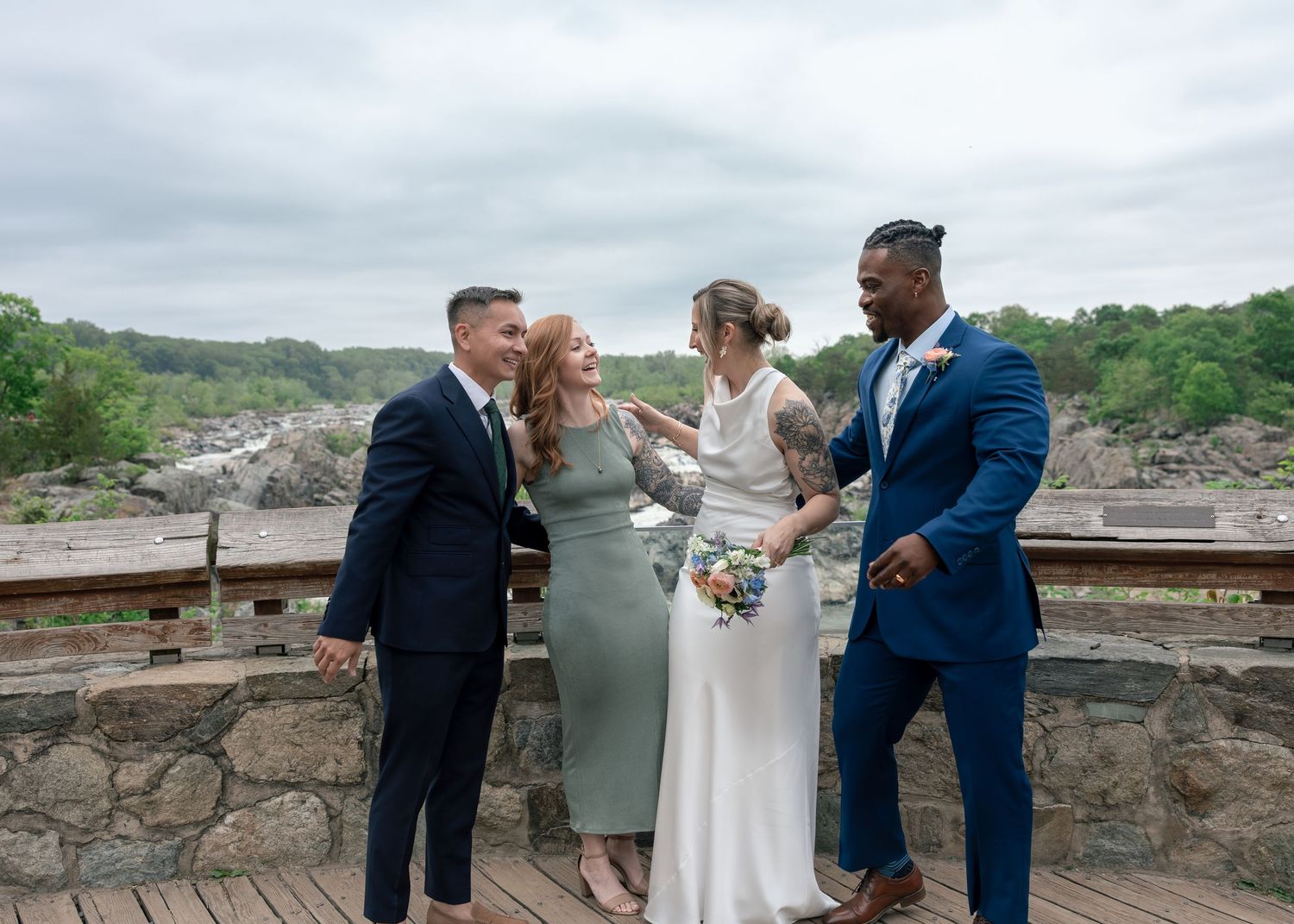 Two couples in formal attire laugh together on a scenic overlook with river views on a wedding day.