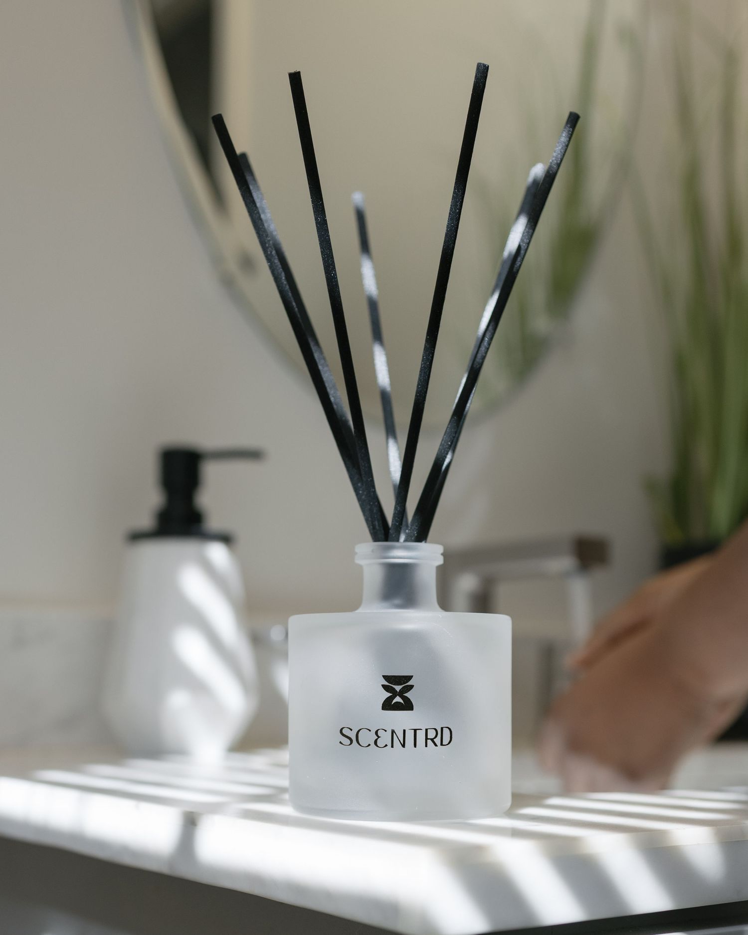Scentrd frosted glass reed diffuser with black reeds on a bathroom shelf beside a white soap dispenser.