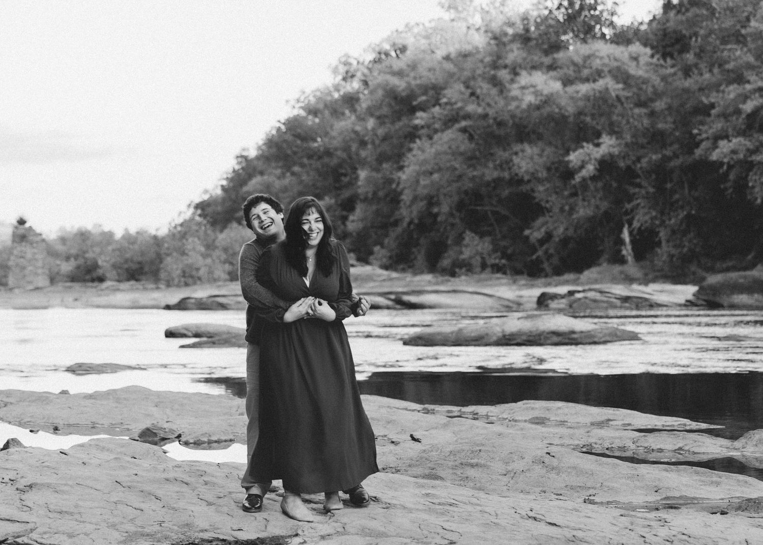 Black and white panoramic photograph showing people walking along a rocky riverbank with trees in the background.