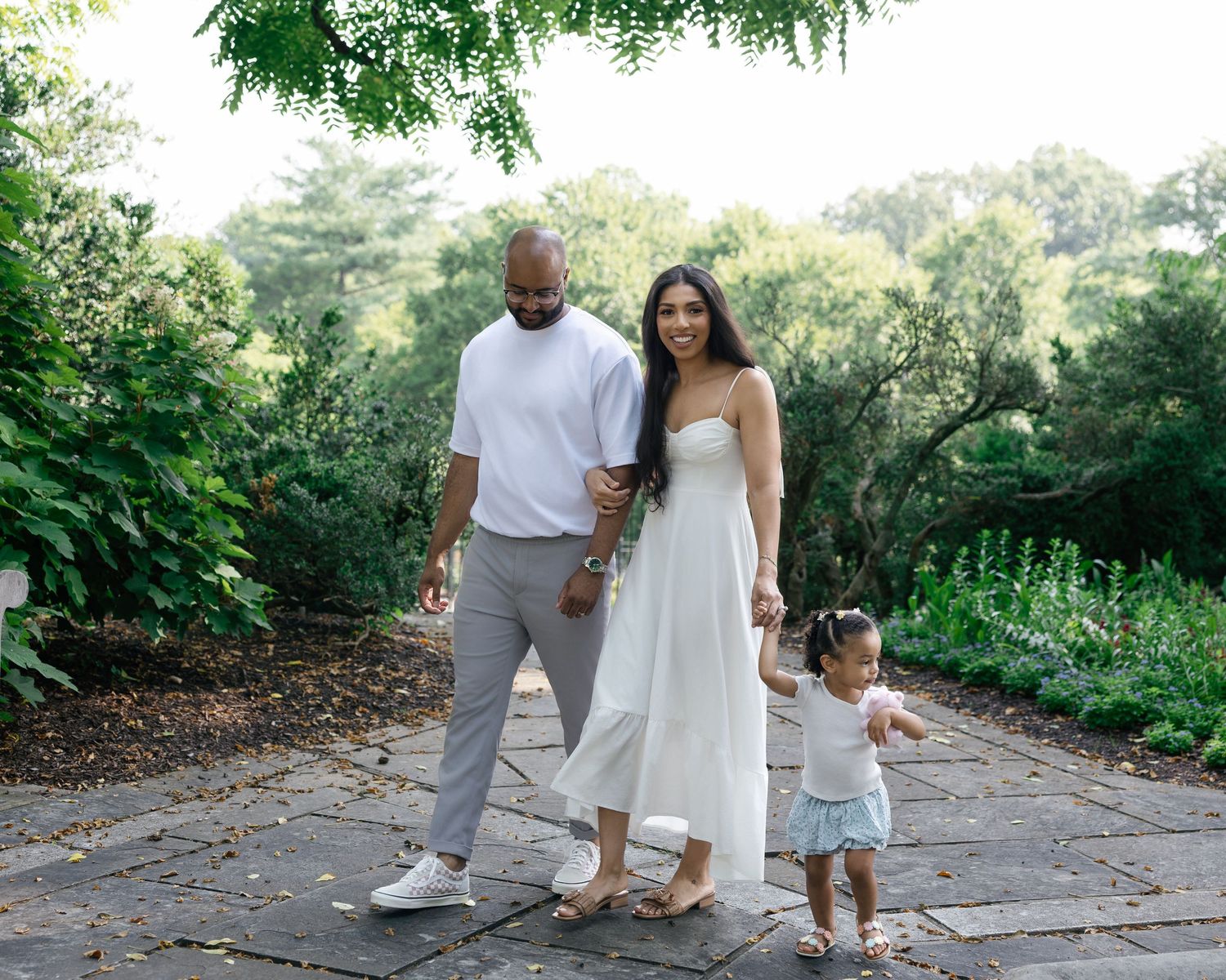 A family of three poses on a garden path, parents holding hands with their toddler daughter, surrounded by lush greenery.