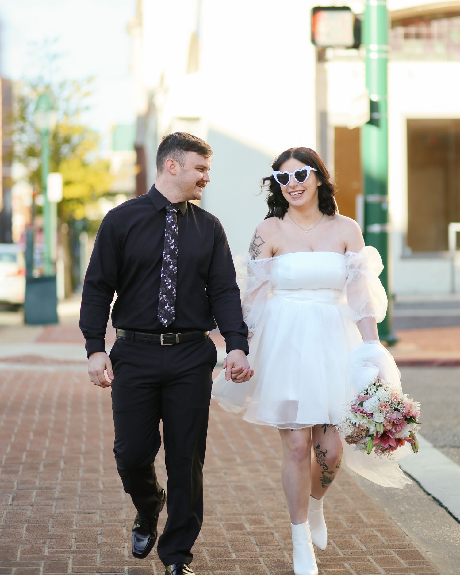 A couple walks hand in hand on brick pavement, with one wearing a black suit and the other in a white dress and sunglasses.