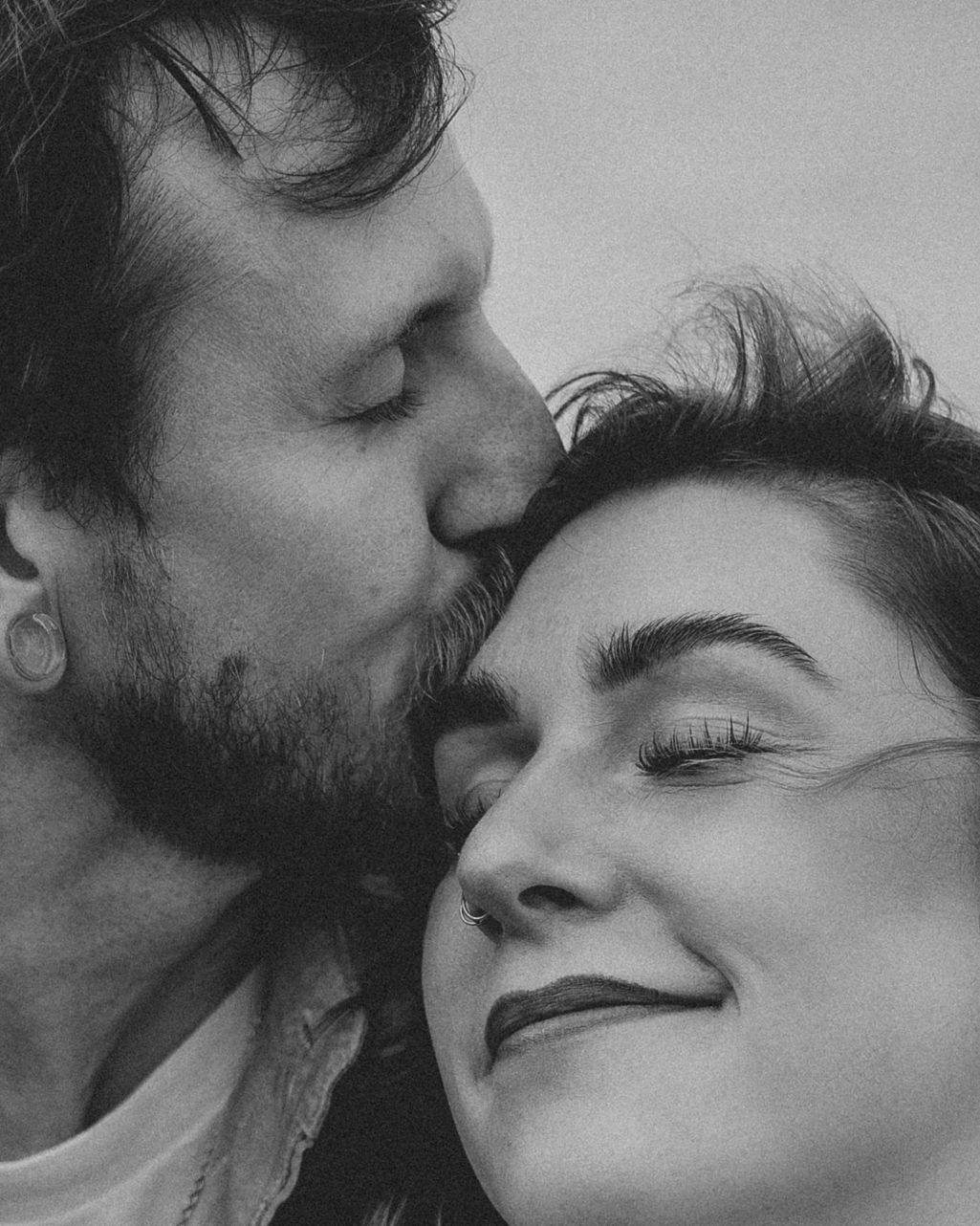 Black and white photo of a bearded man kissing a smiling woman on the cheek in a tender romantic moment.