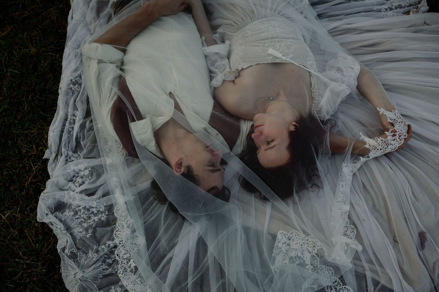 Two people in elegant white lace dresses lying on grass with delicate tulle fabric surrounding them.