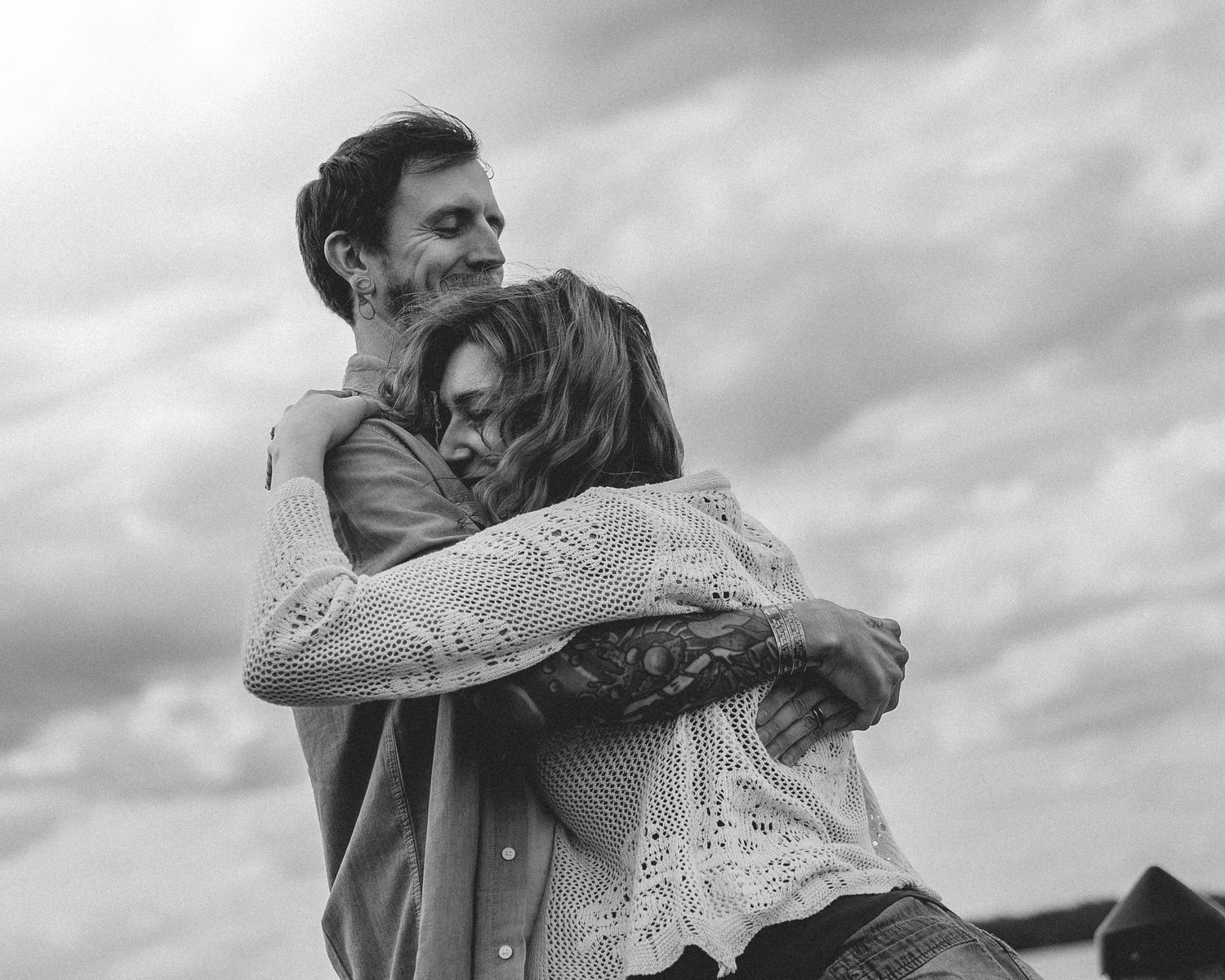 Black and white photo of a couple embracing tenderly outdoors under a cloudy sky.
