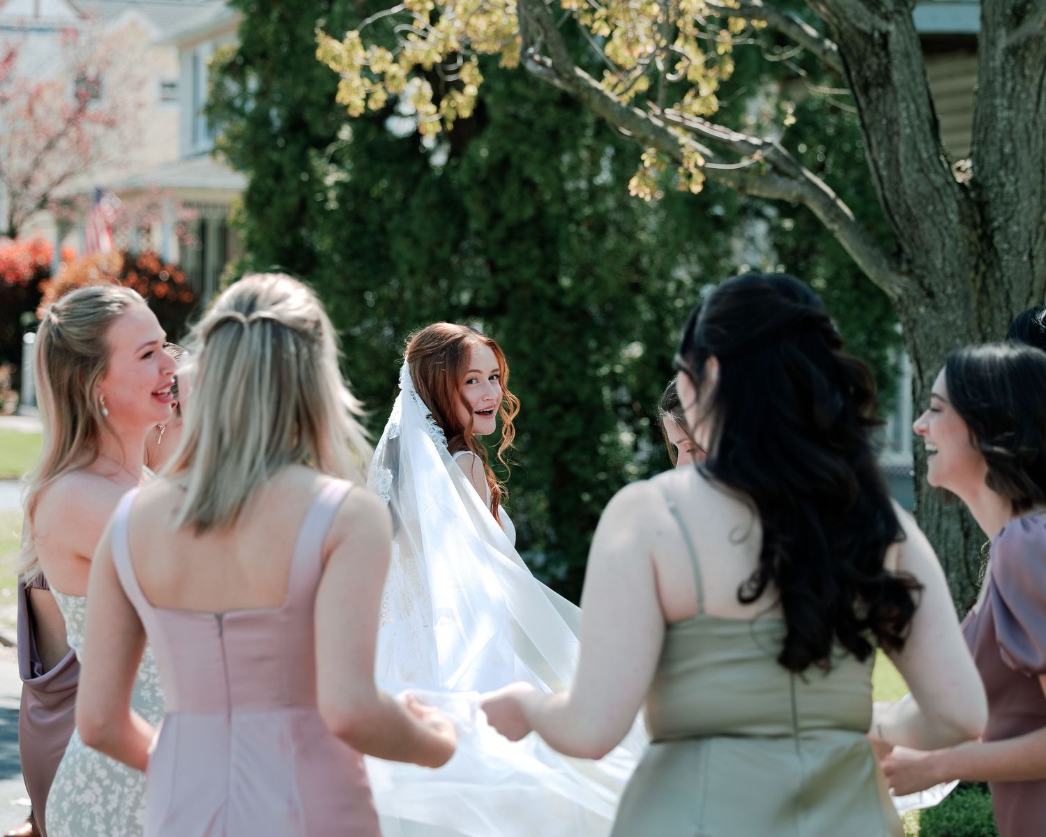 Bride laughing with bridesmaids outdoors, veil flowing, surrounded by spring blossoms at a wedding celebration.