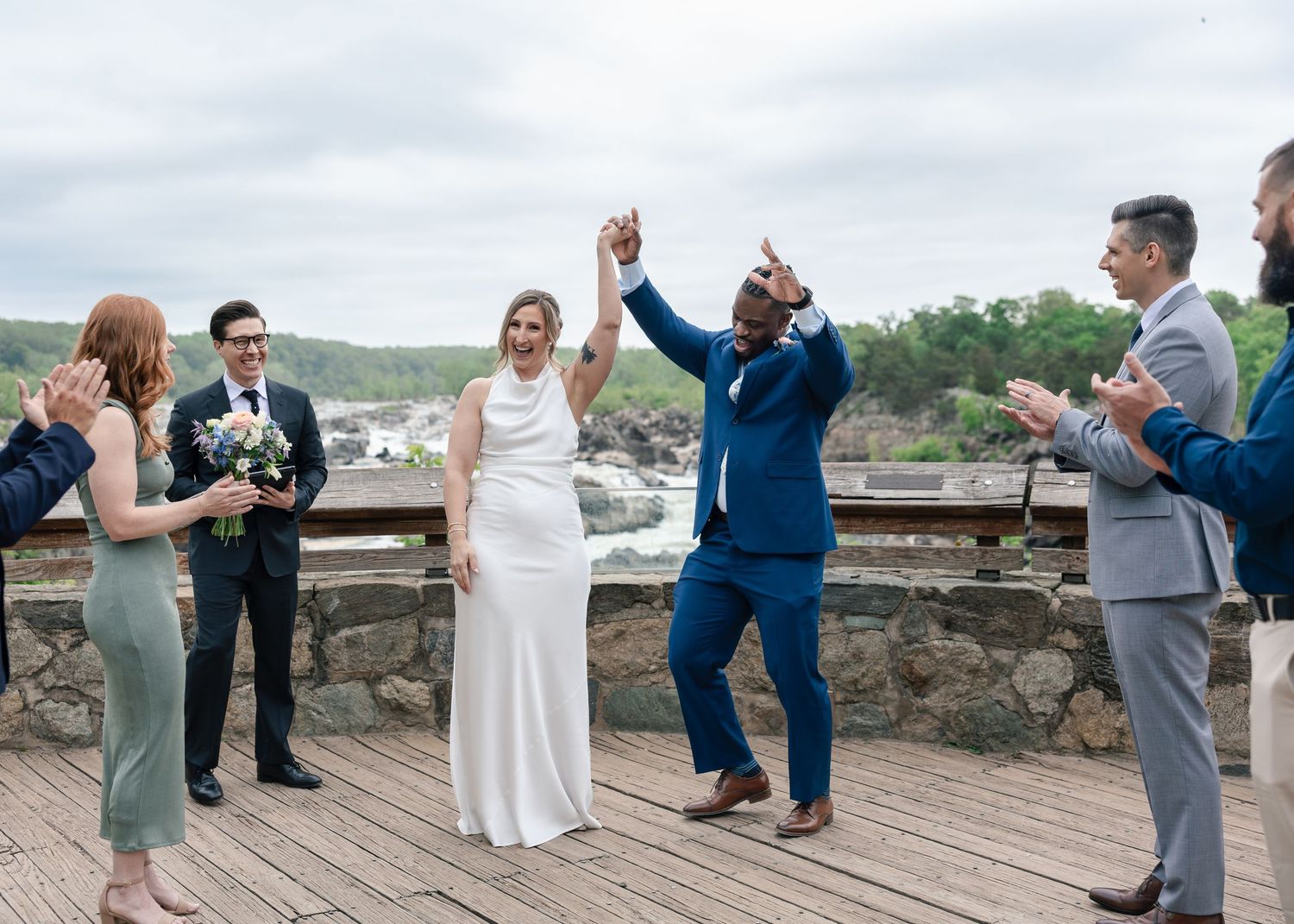 Joyful newlyweds celebrate after wedding ceremony outdoors, groom raises arms in triumph, guests applaud on scenic overlook.