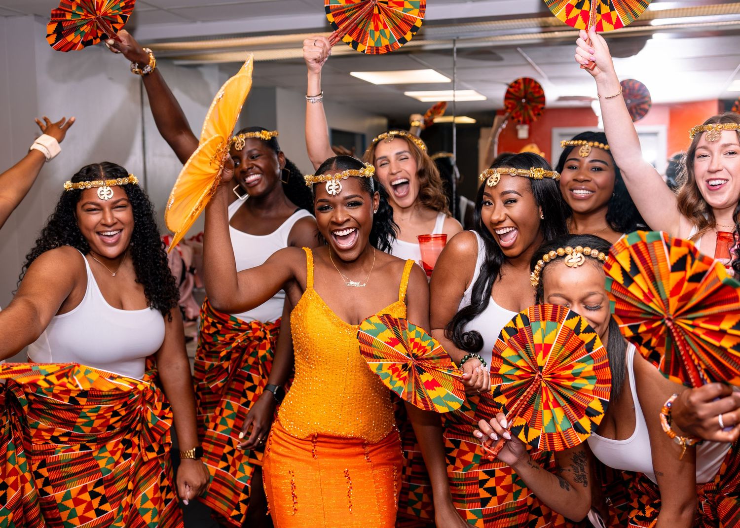 Group of women in colorful Kente cloth outfits celebrating together, holding decorative fans at a festive cultural event.