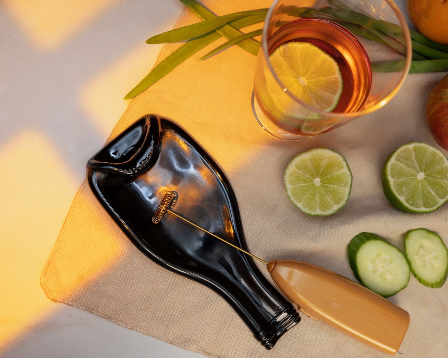 Black flattened wine bottle spoon rest beside sliced limes, cucumber, and a glass of iced tea on a linen surface.