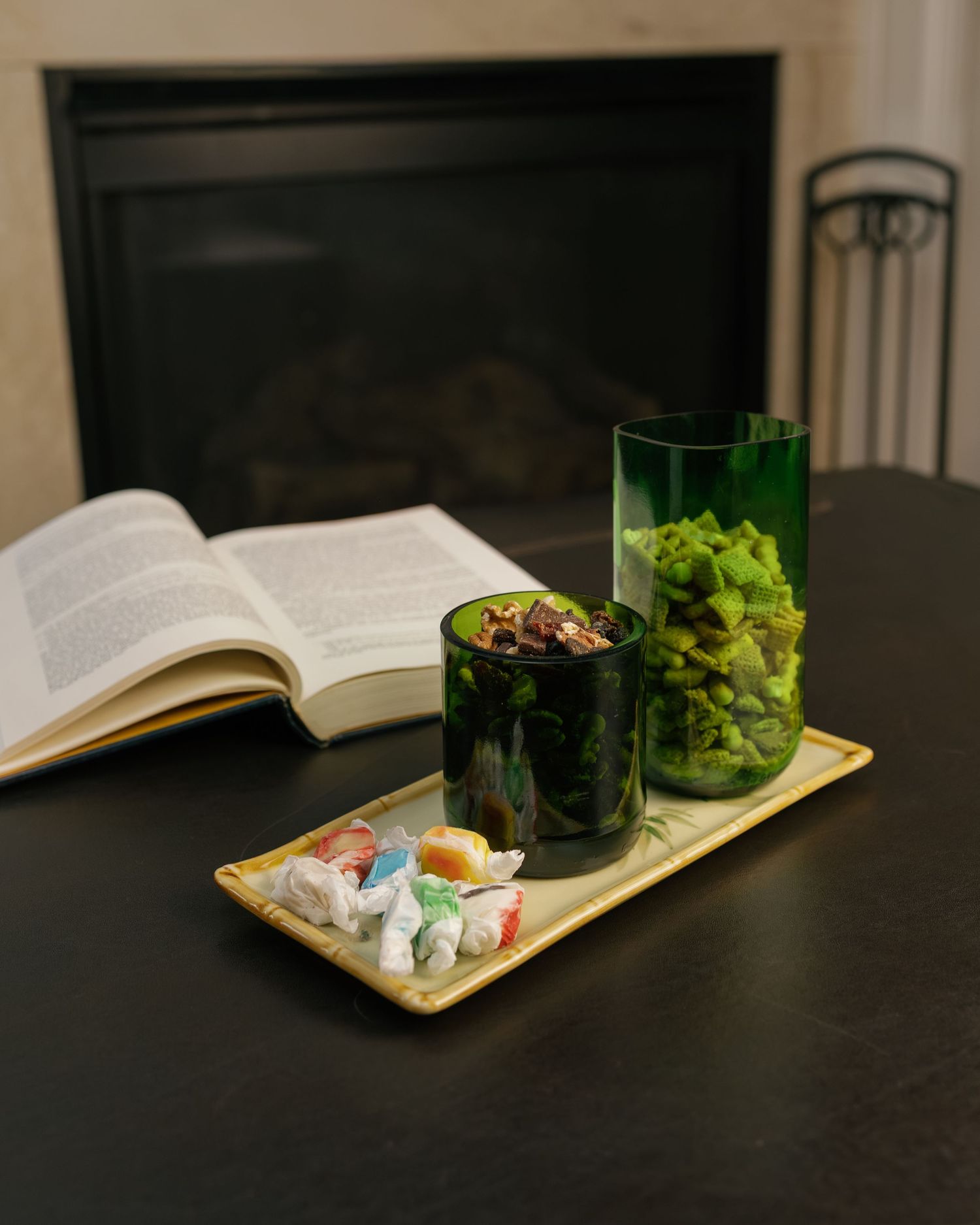 Green glass vases with plants, colorful taffy candy on a tray, and an open book near a fireplace on a dark table.