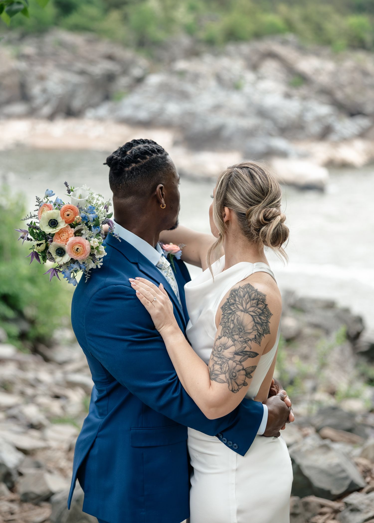 Couple embracing outdoors on rocky riverbank, groom in navy suit holding bouquet, bride in white dress with floral shoulder tattoo.