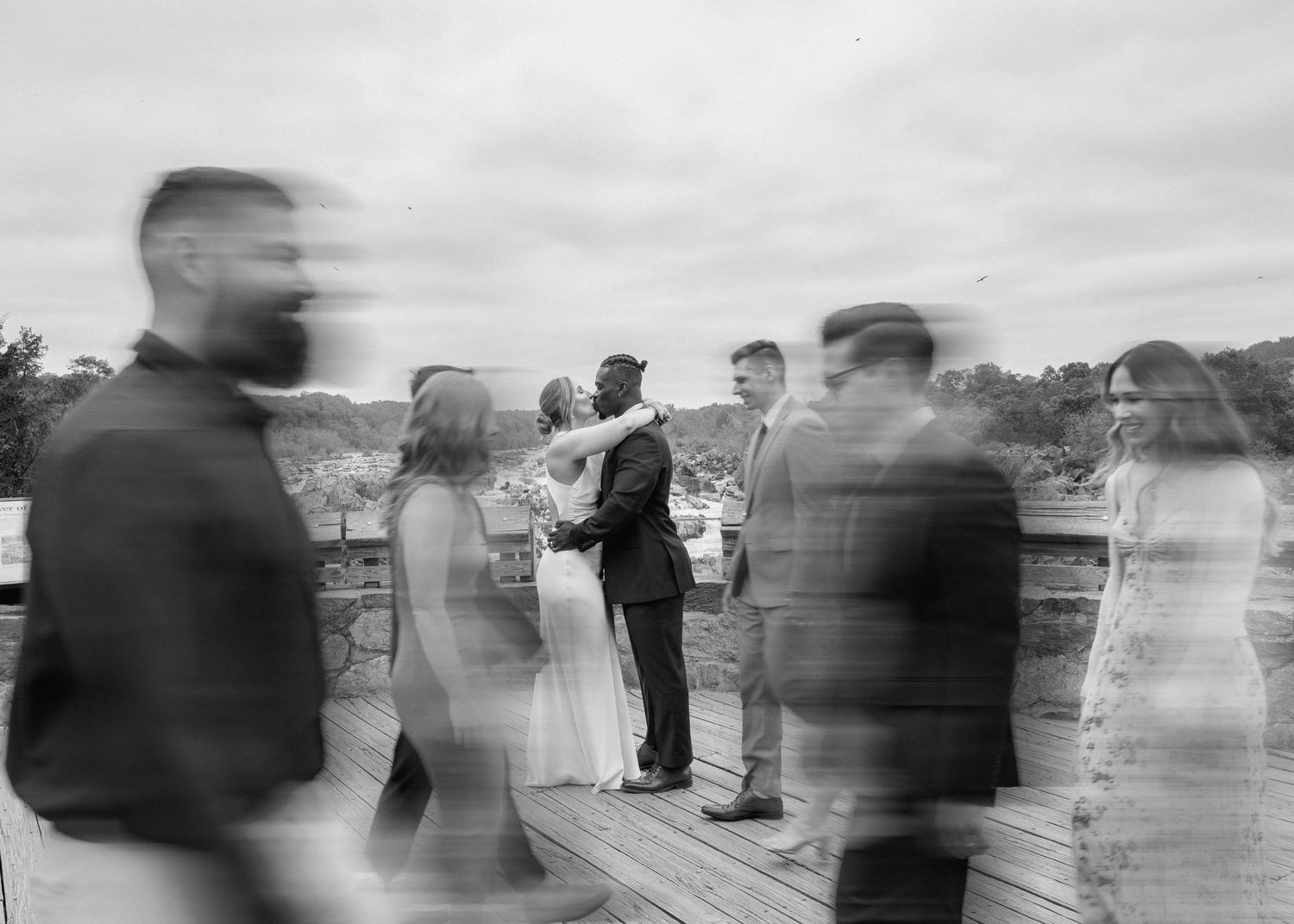 Blurred wedding guests surround a couple sharing a romantic moment on a wooden deck in a dreamy black and white photo.