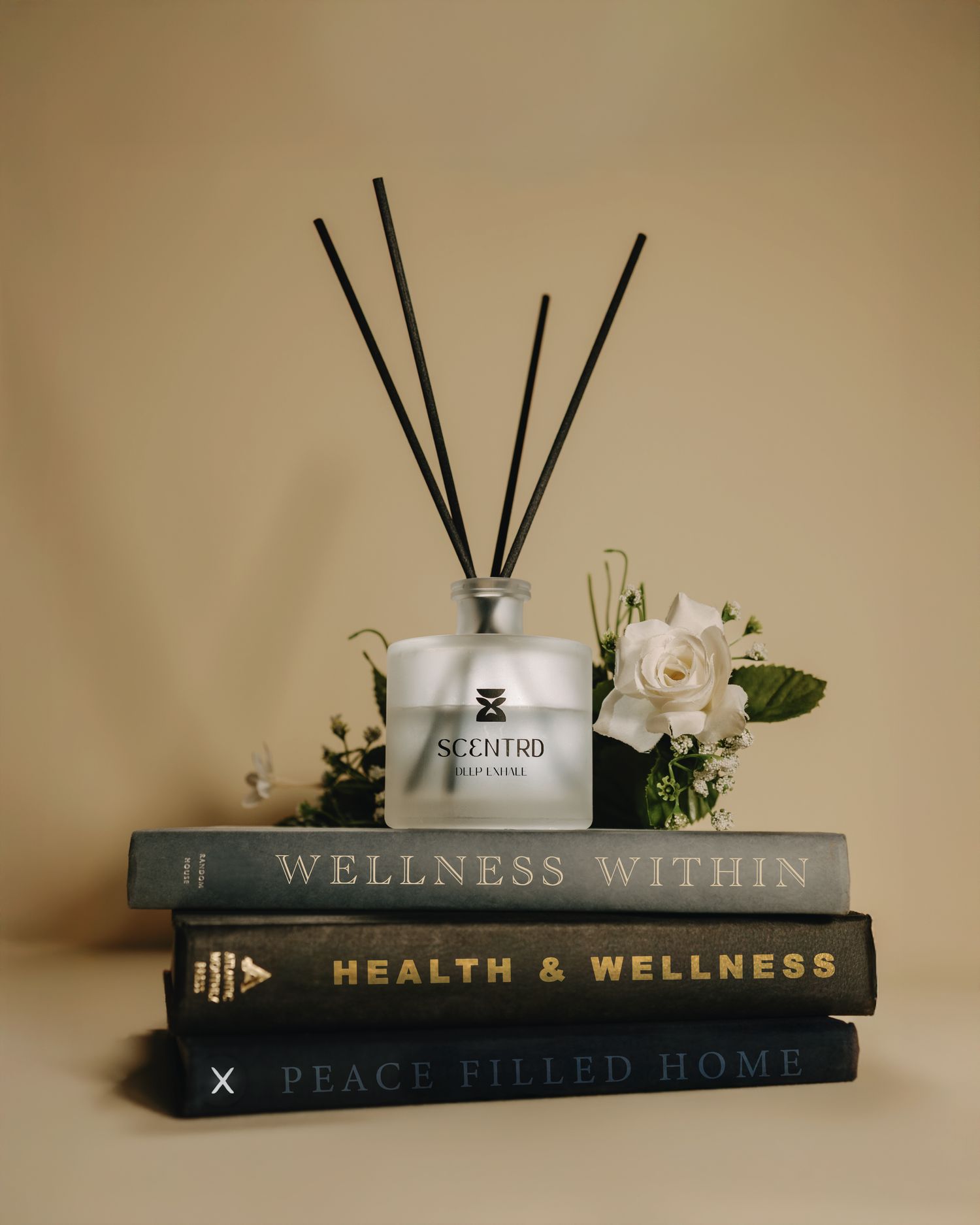 A SCENTRD reed diffuser sits atop stacked wellness books with white roses on a beige background.