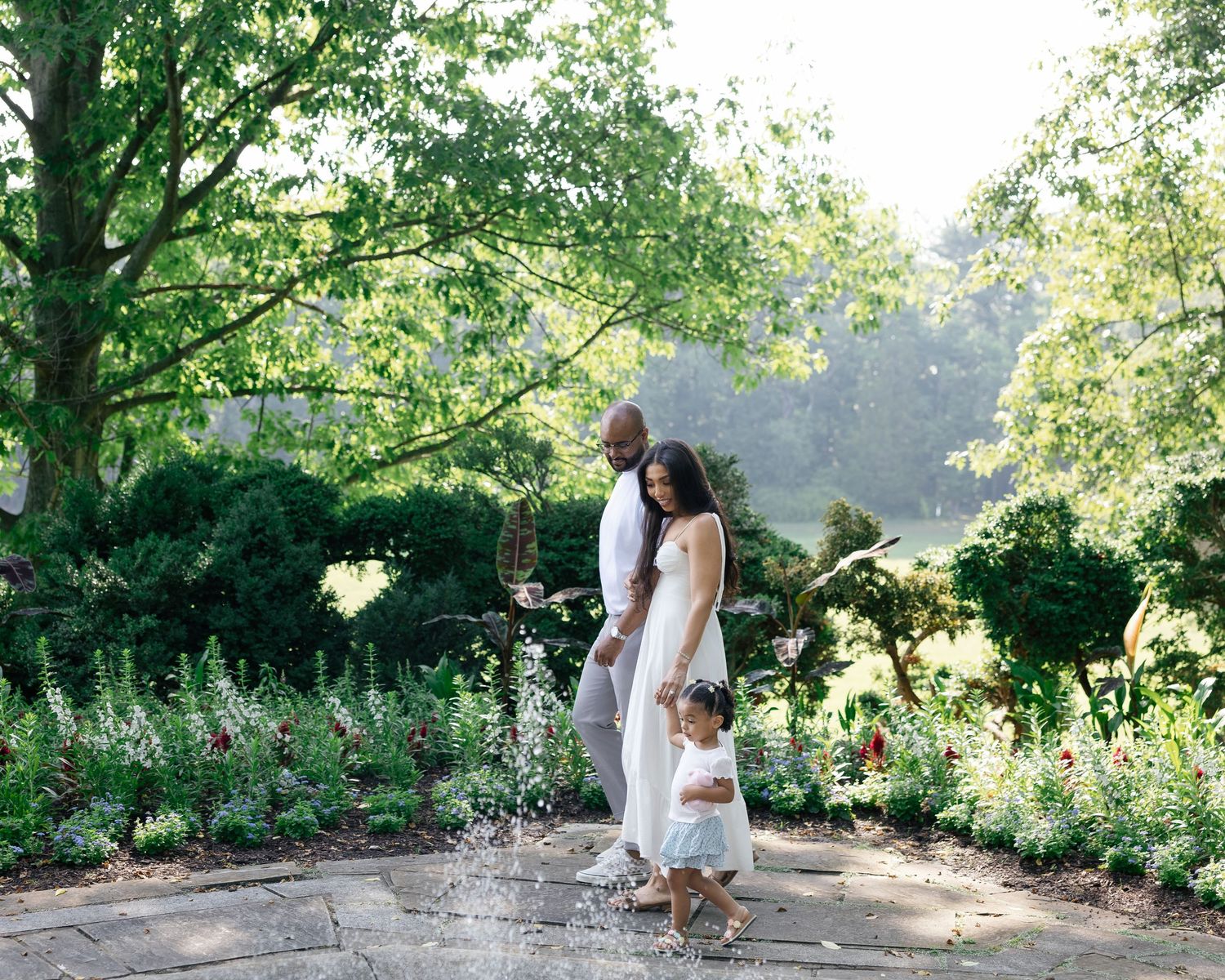 Family in white outfits posing by a garden fountain surrounded by lush greenery and colorful flowers.
