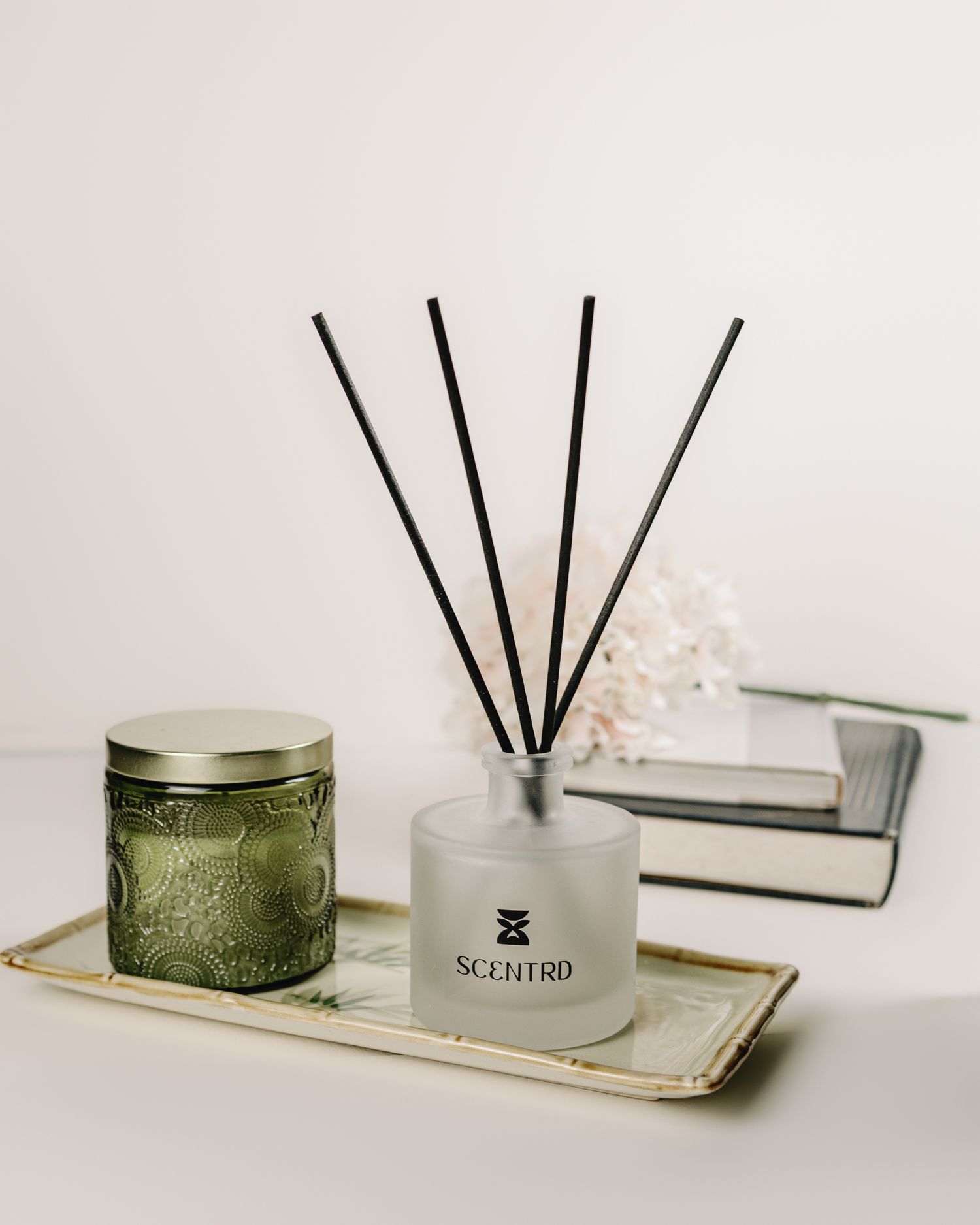 Scentrd reed diffuser and green embossed candle tin on decorative tray with books and coral accent.