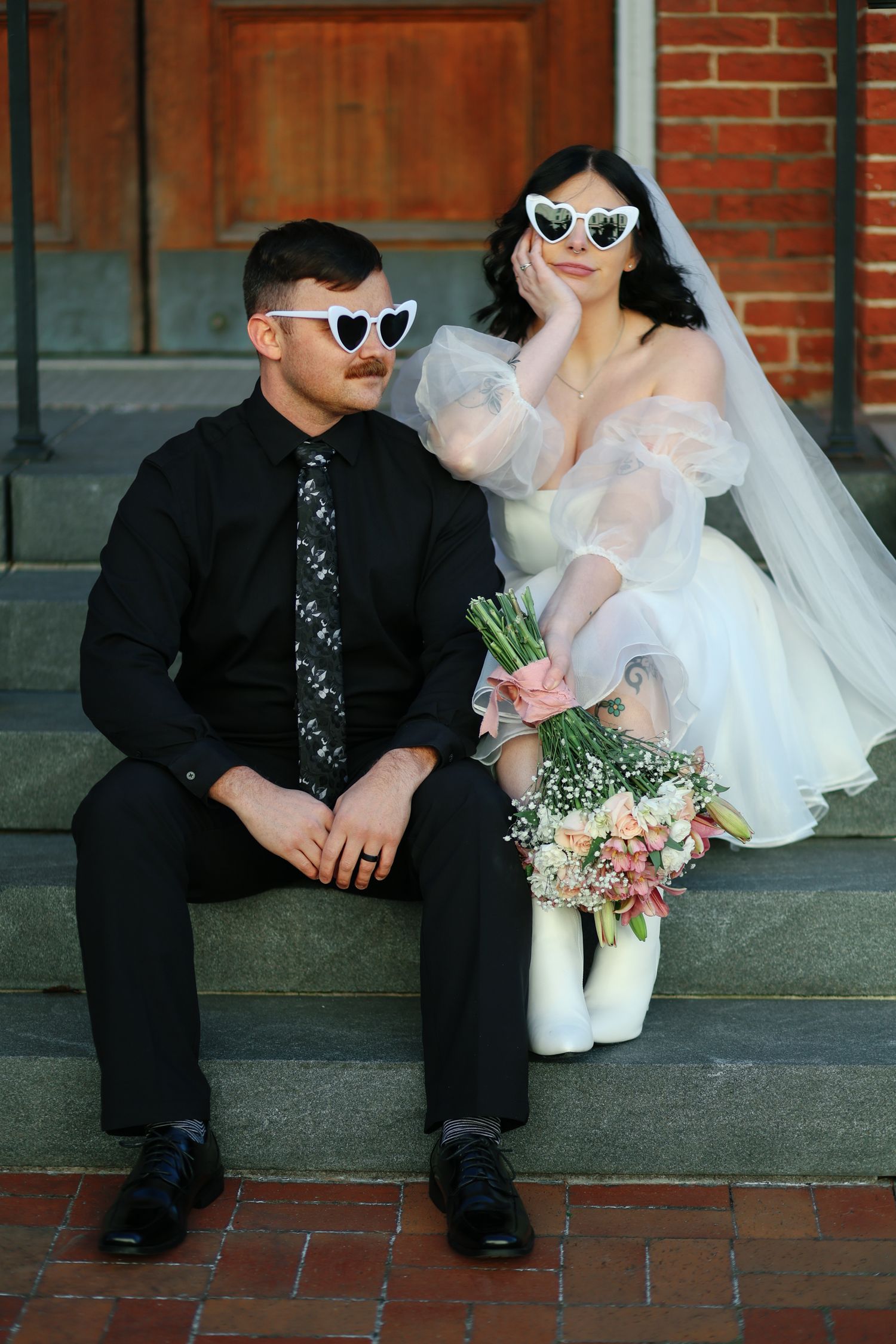 Wedding couple sits together on steps with bride in white dress holding pink flowers and both wearing sunglasses.