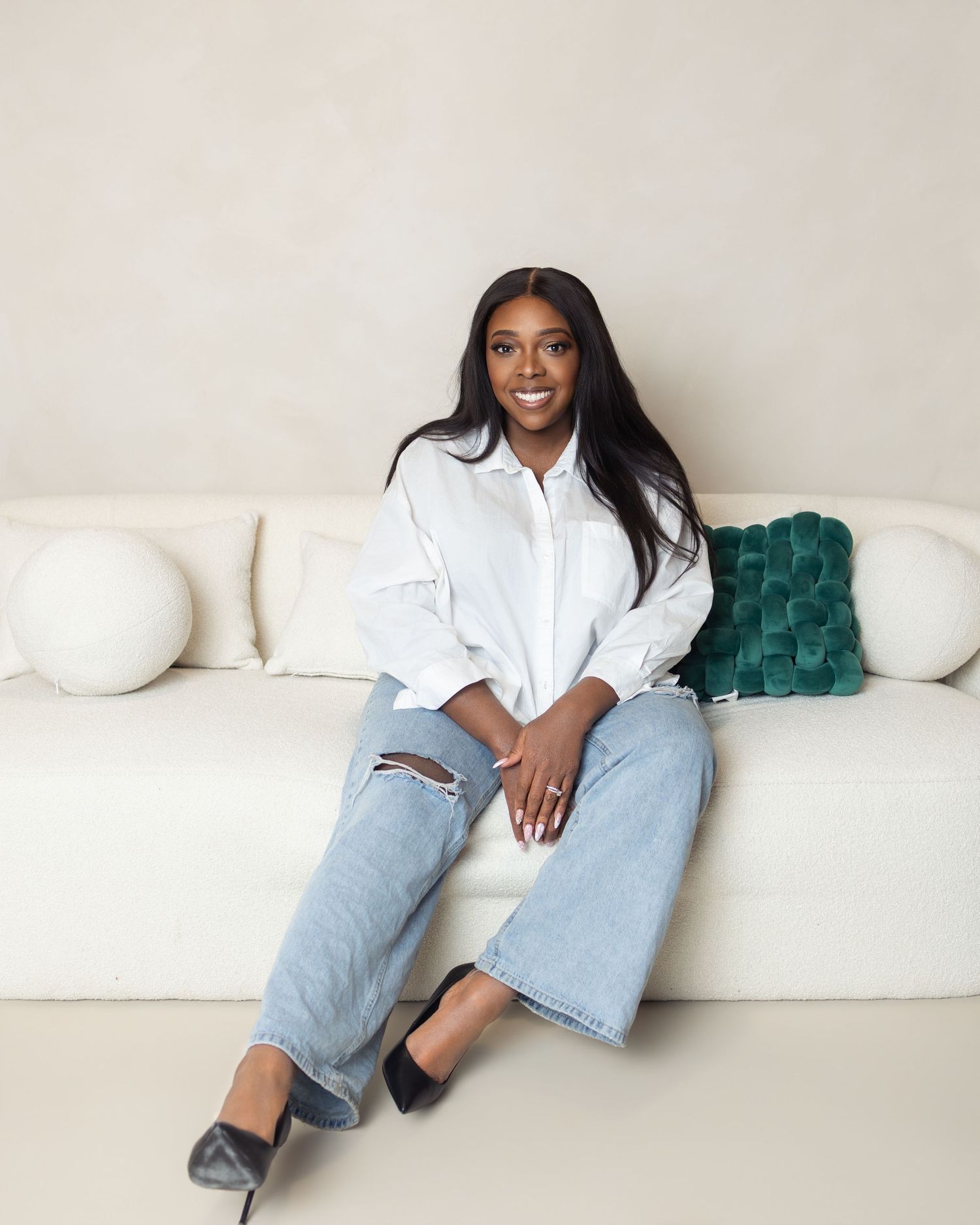 Young woman in white shirt and ripped jeans sitting on a white sofa with a green velvet pillow.
