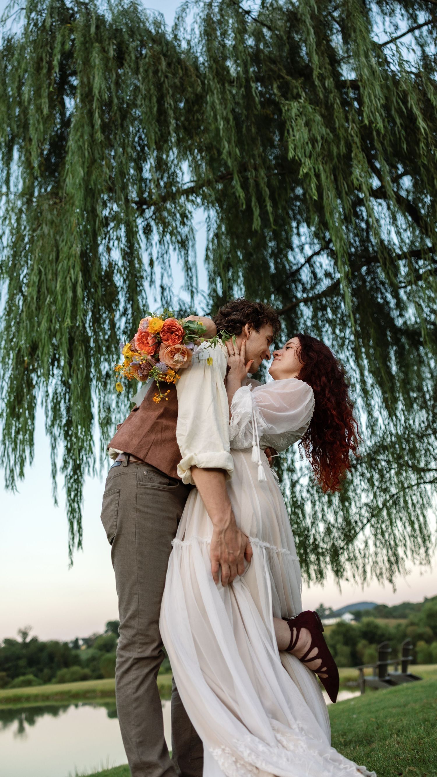 Romantic couple in wedding attire dances and embraces under a weeping willow tree at sunset by a lake.